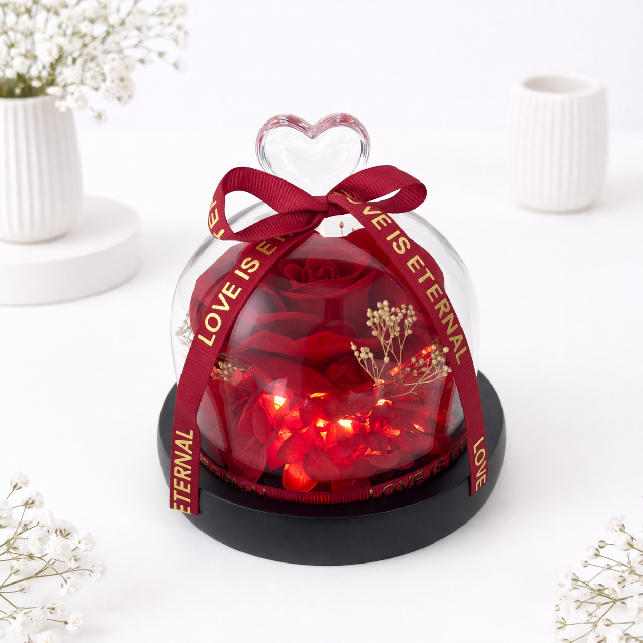 A preserved red rose with LED lights inside a glass dome, topped with a heart handle and "Love Is Eternal" ribbon.
