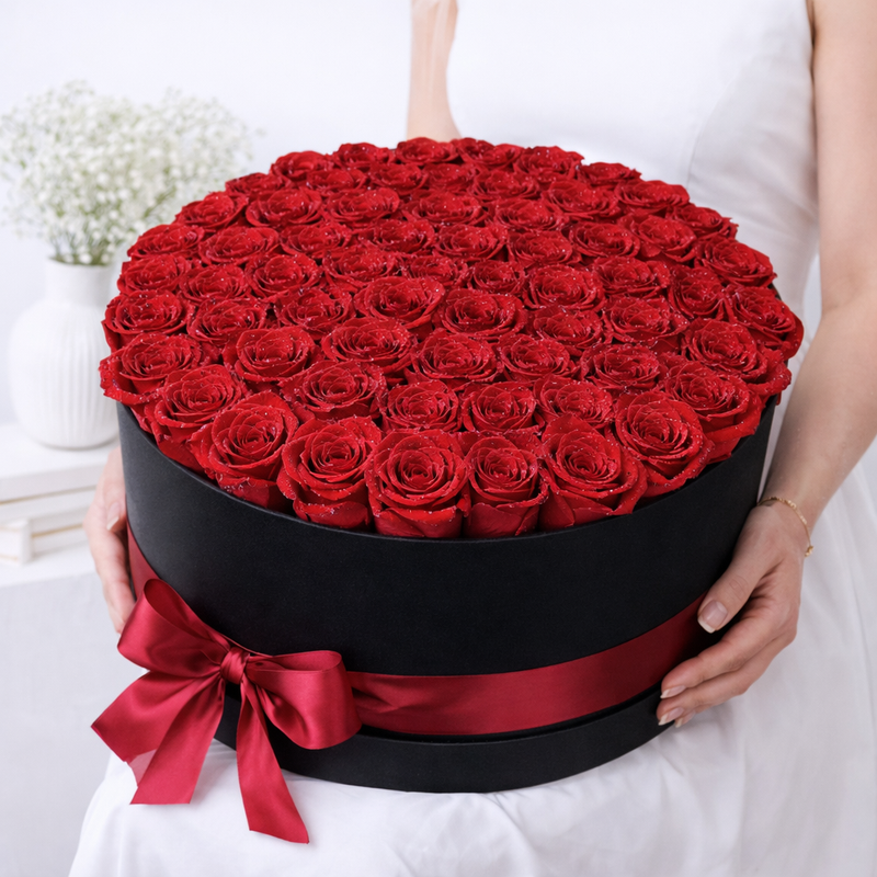 A woman holding a large round black box filled with red glitter roses, tied with a red satin ribbon.