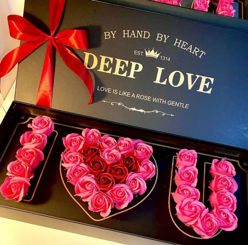Pink and red soap roses arranged in "I ♥ U" inside an open black "Deep Love" box with a red ribbon bow.