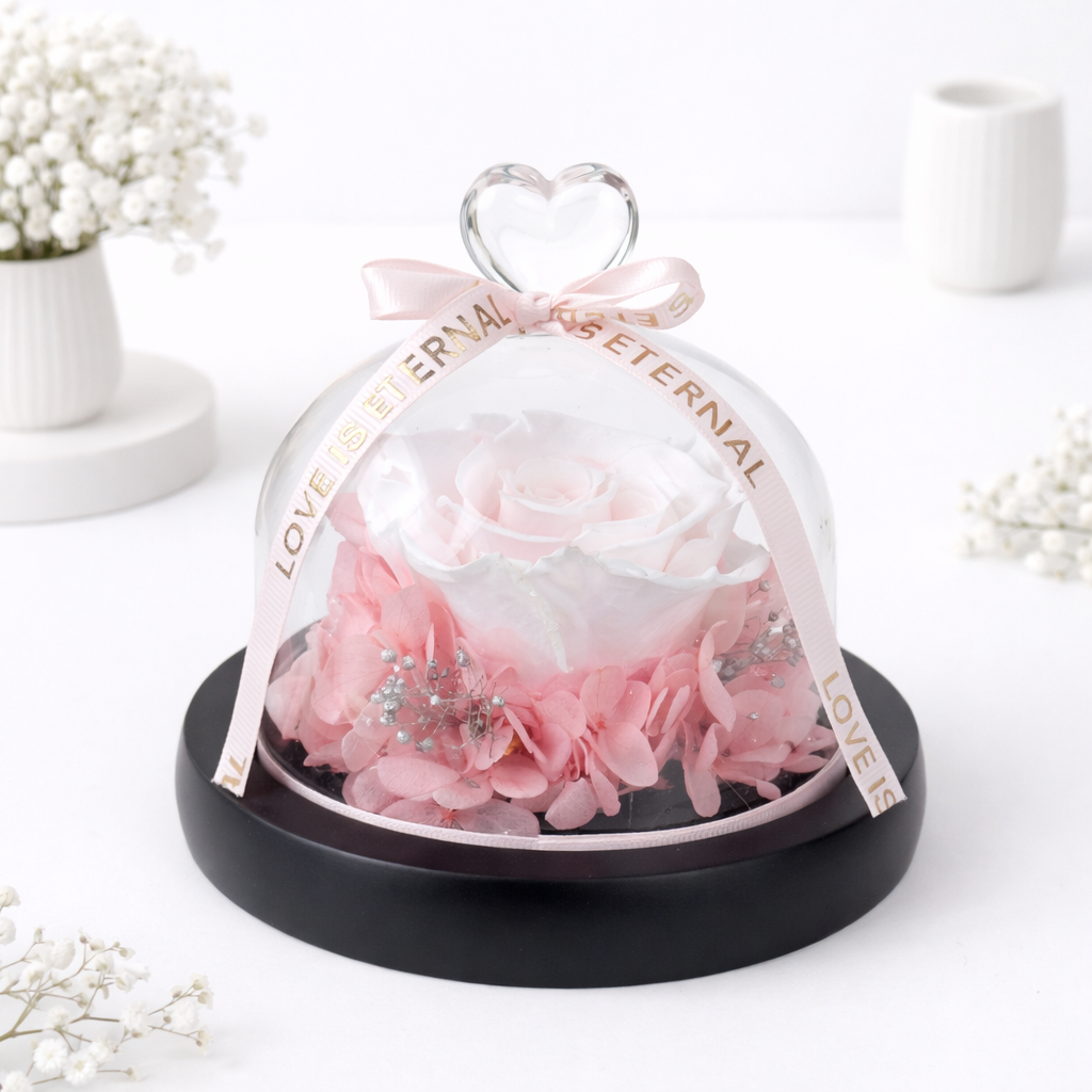 Pink preserved rose with hydrangeas under a glass dome with a crystal heart handle and "Love Is Eternal" ribbon on a black base.