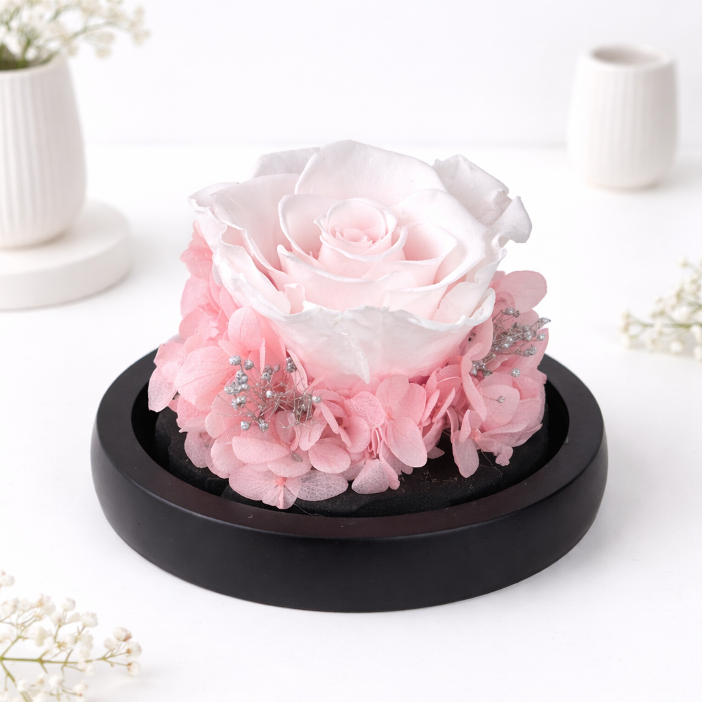 Close-up of a single pink preserved rose surrounded by hydrangeas and silver baby's breath on a black wooden base.