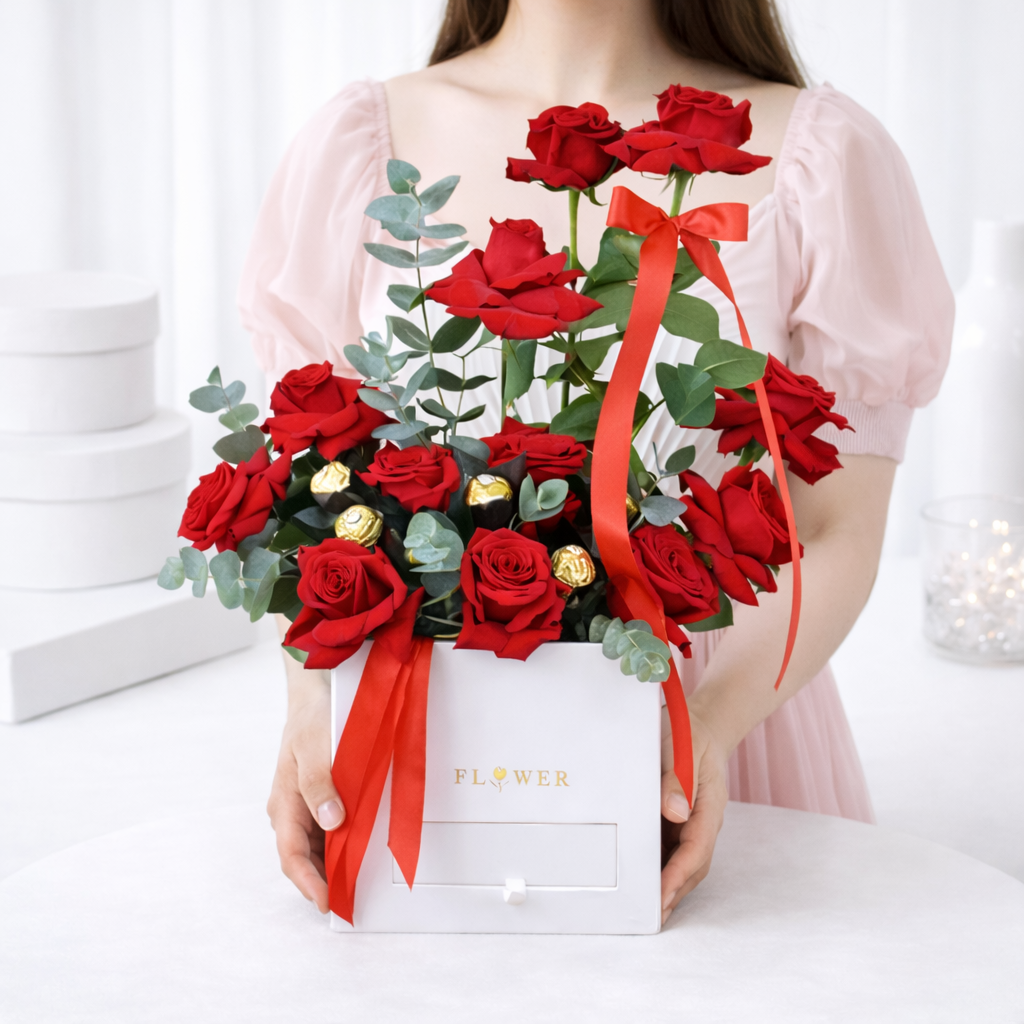 Woman in a pink dress holding a white gift box of red roses, Ferrero Rocher chocolates, and eucalyptus tied with a red ribbon.