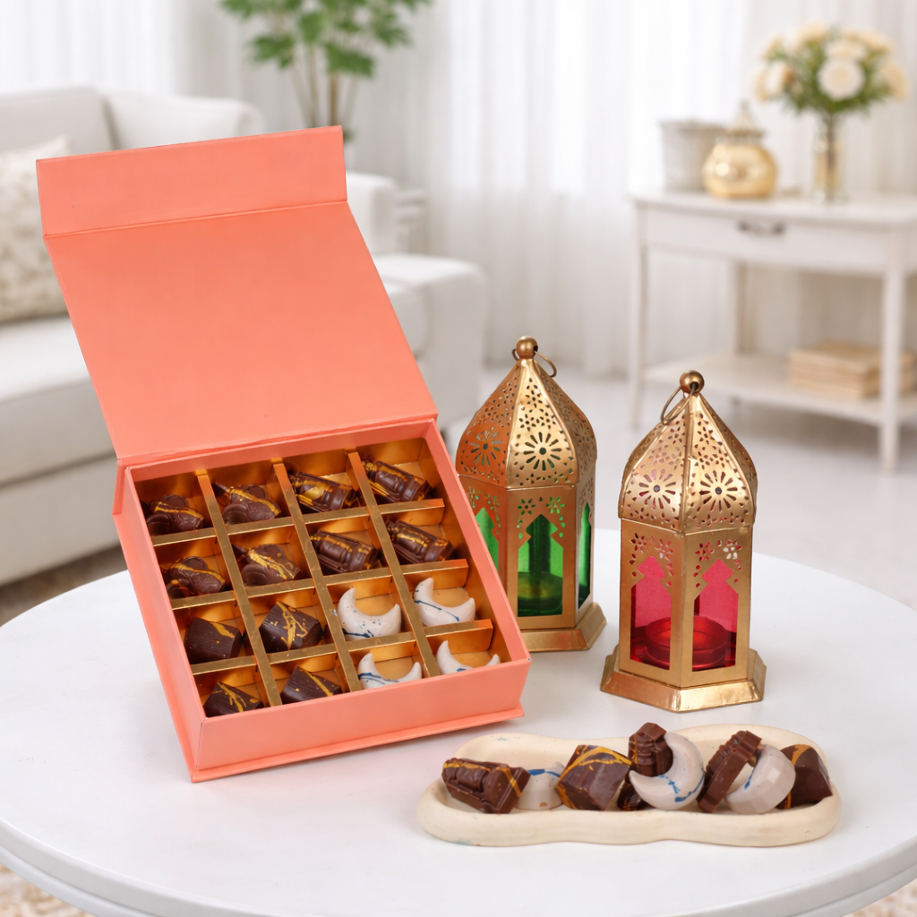 An orange gift box showcases 16 artisanal chocolates in a gold grid, featuring shapes like dark blue moons, light blue domes, and green lanterns. The set is styled on a white table with two gold lanterns and a marble tray holding loose chocolates.
