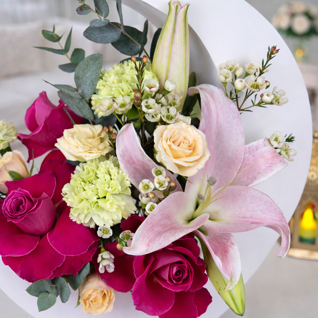 A close-up view of the moon floral arrangement, highlighting the textures of the pink lily petals, the deep magenta roses, and the small white waxflower accents nestled among eucalyptus leaves.