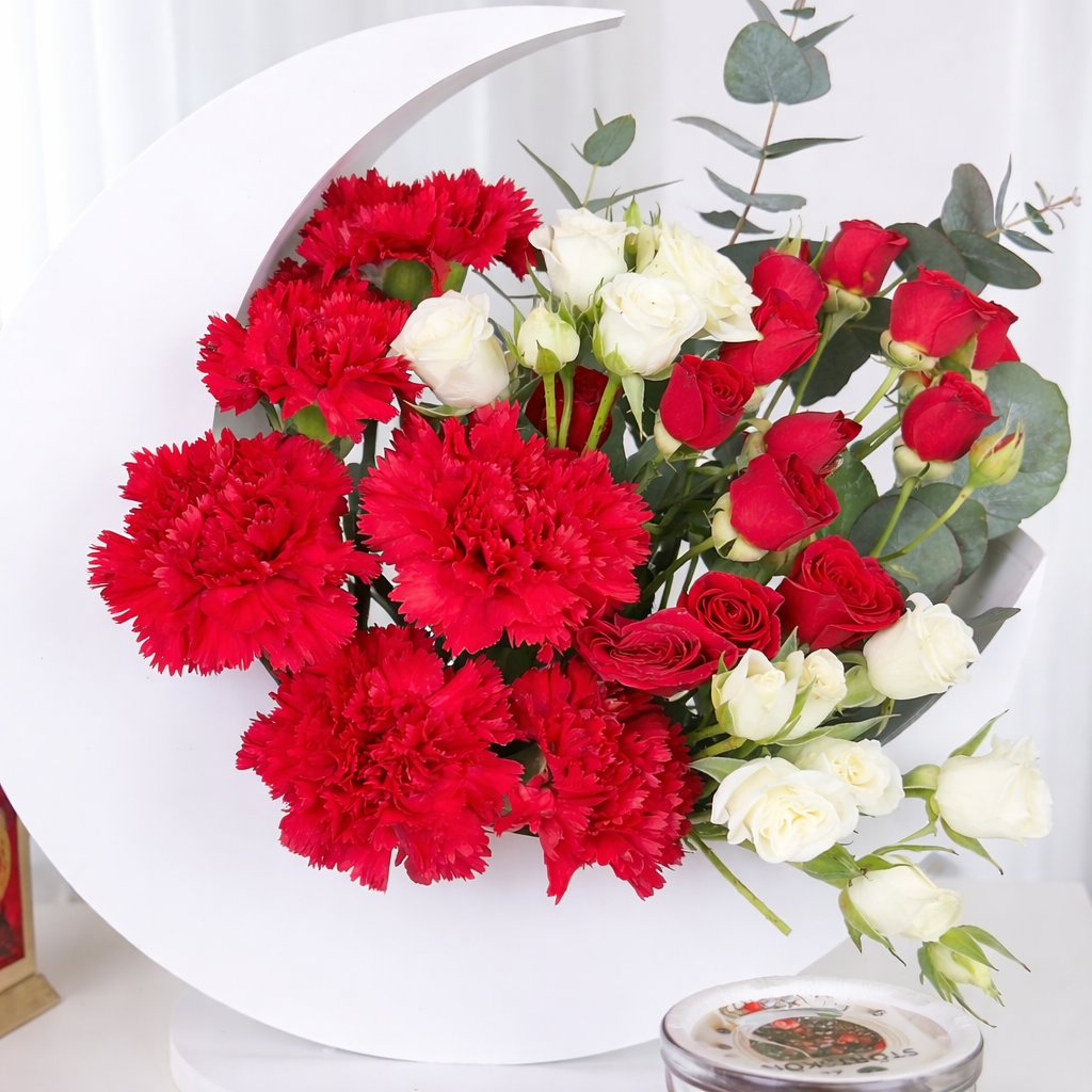 A full display of a white crescent moon flower stand filled with vibrant red carnations, red rosebuds, and white spray roses, accompanied by a black tasbih, a candle, and a gold lantern.