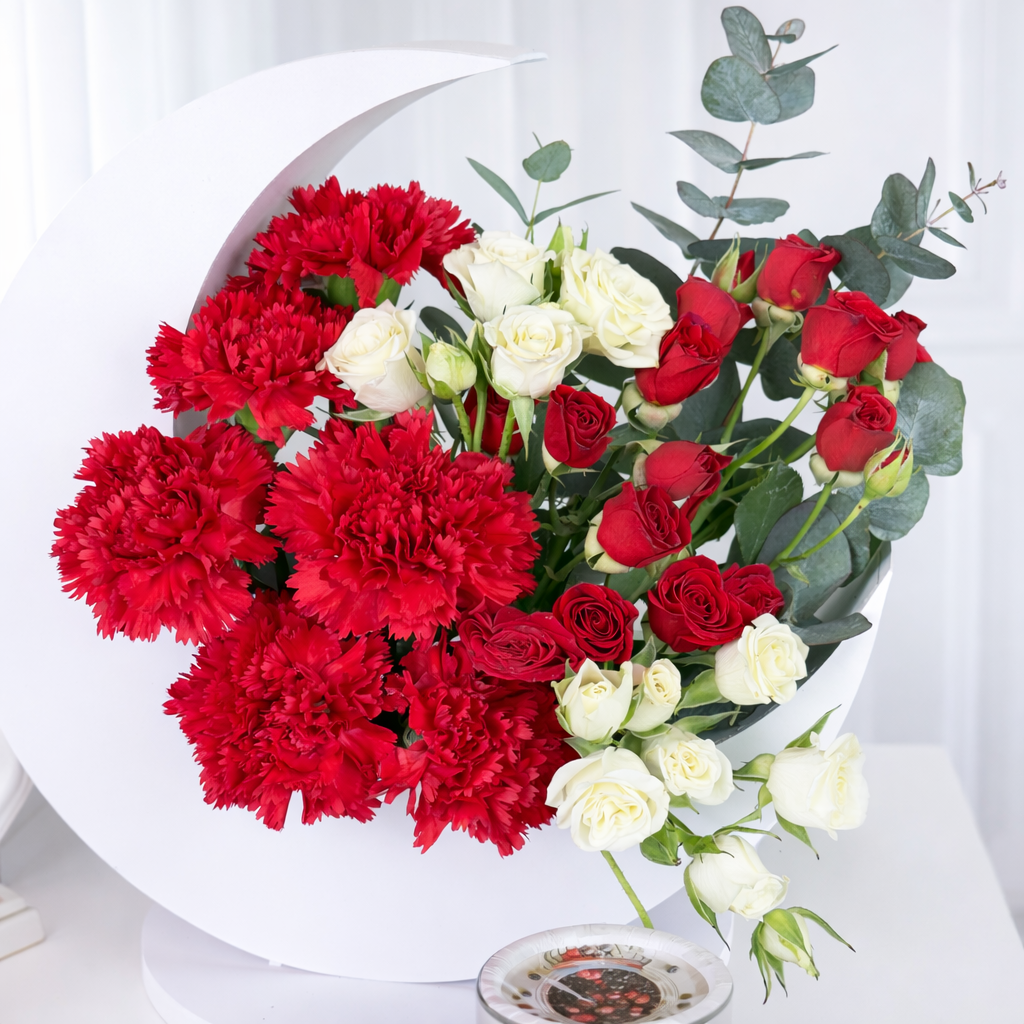 A centered close-up of a festive red and white floral arrangement in a crescent moon frame, emphasizing the contrast between the ruffled red carnations and delicate white rose blossoms.