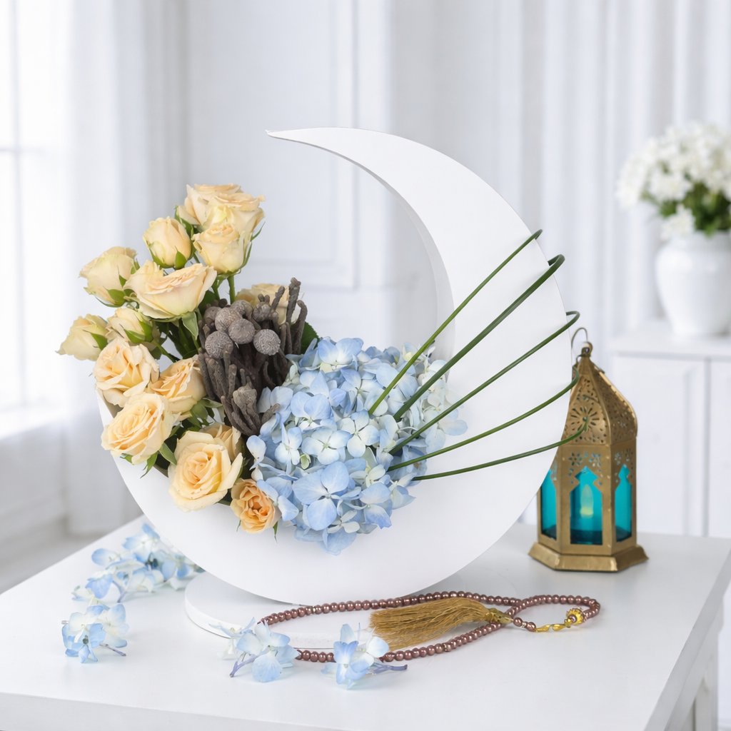 A crescent moon-shaped floral arrangement with peach roses and blue hydrangeas, displayed on a white table with a gold Ramadan lantern and prayer beads.