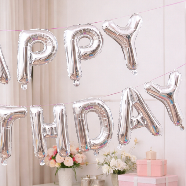 Close-up of silver 'Happy Birthday' foil letter balloons with flowers and pink gift boxes in the background.