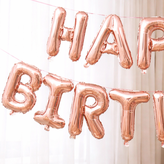 Close-up of rose gold 'Happy Birthday' foil letter balloons on a string against a white curtain.