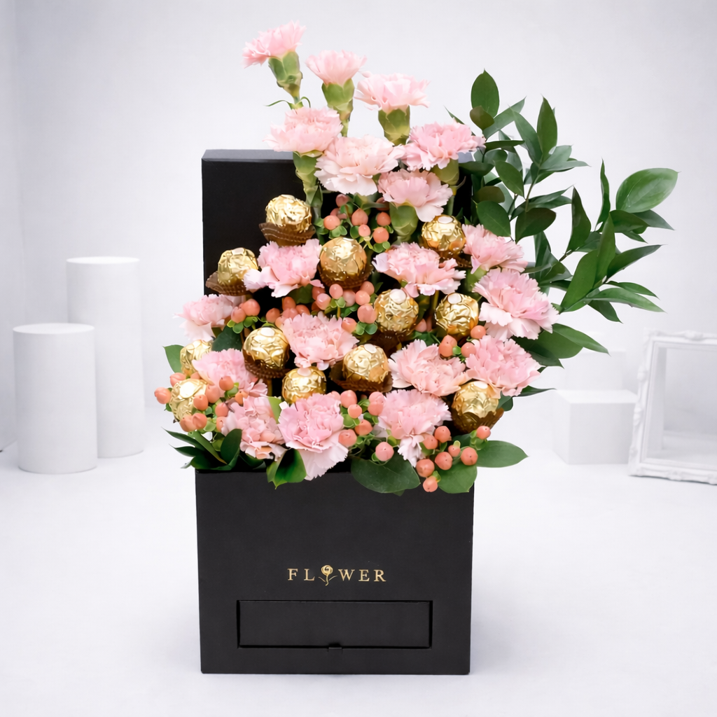 `Pink carnations and Ferrero Rocher chocolates arranged in a black gift box with green foliage.`