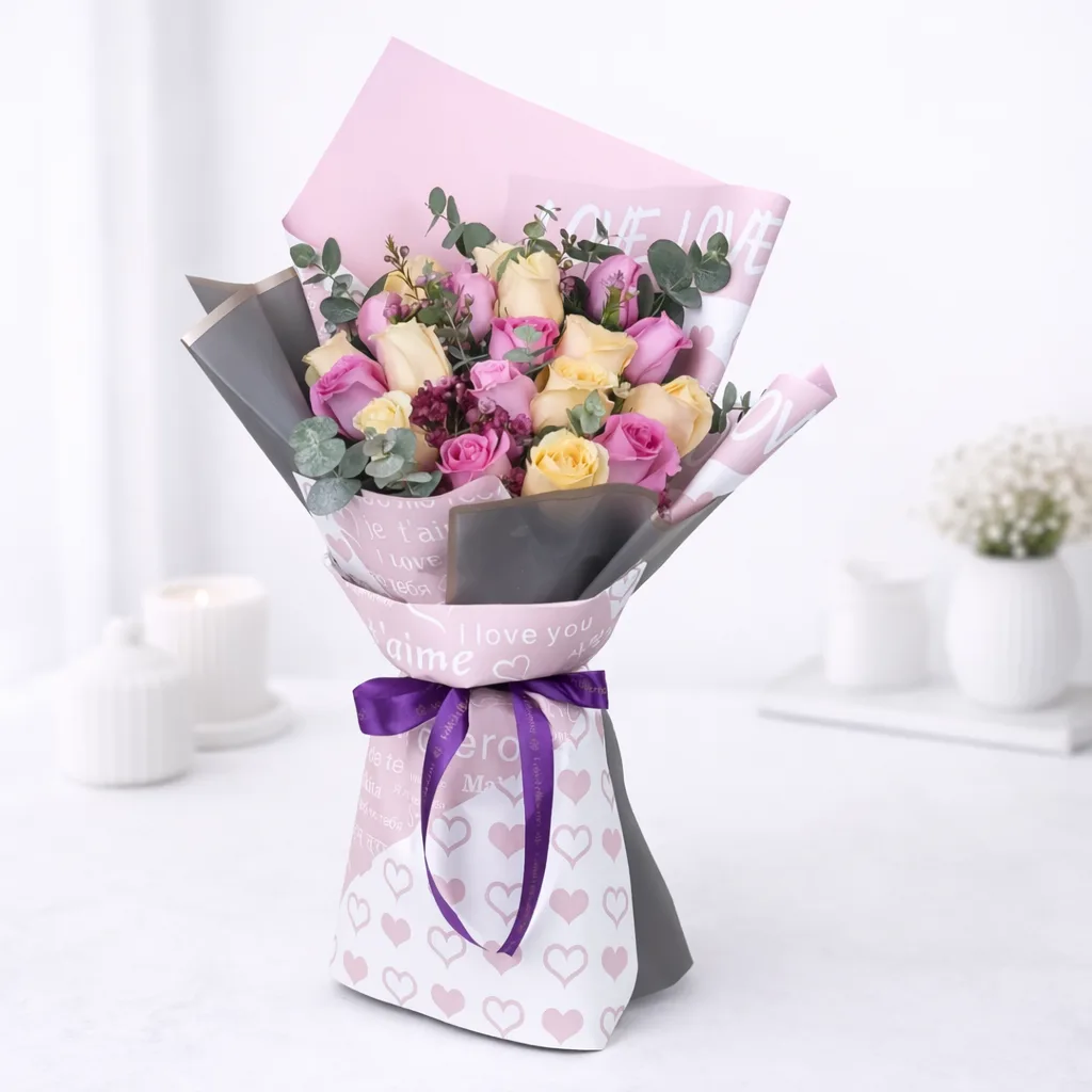 Mixed pink and cream rose bouquet wrapped in "I Love You" printed paper with a purple ribbon bow
