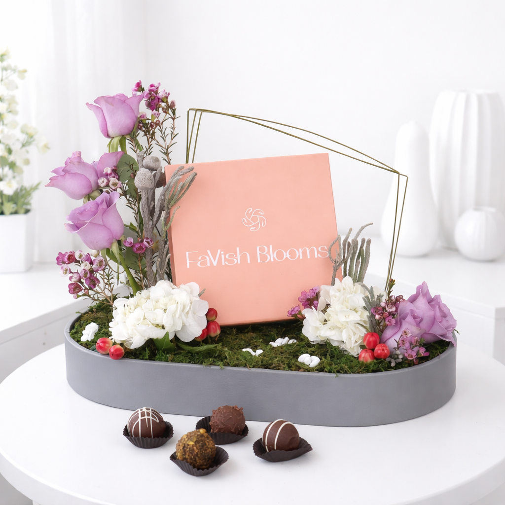 FaVish Blooms floral arrangement tray with purple roses, white hydrangeas, dried flowers, and assorted chocolate truffles.