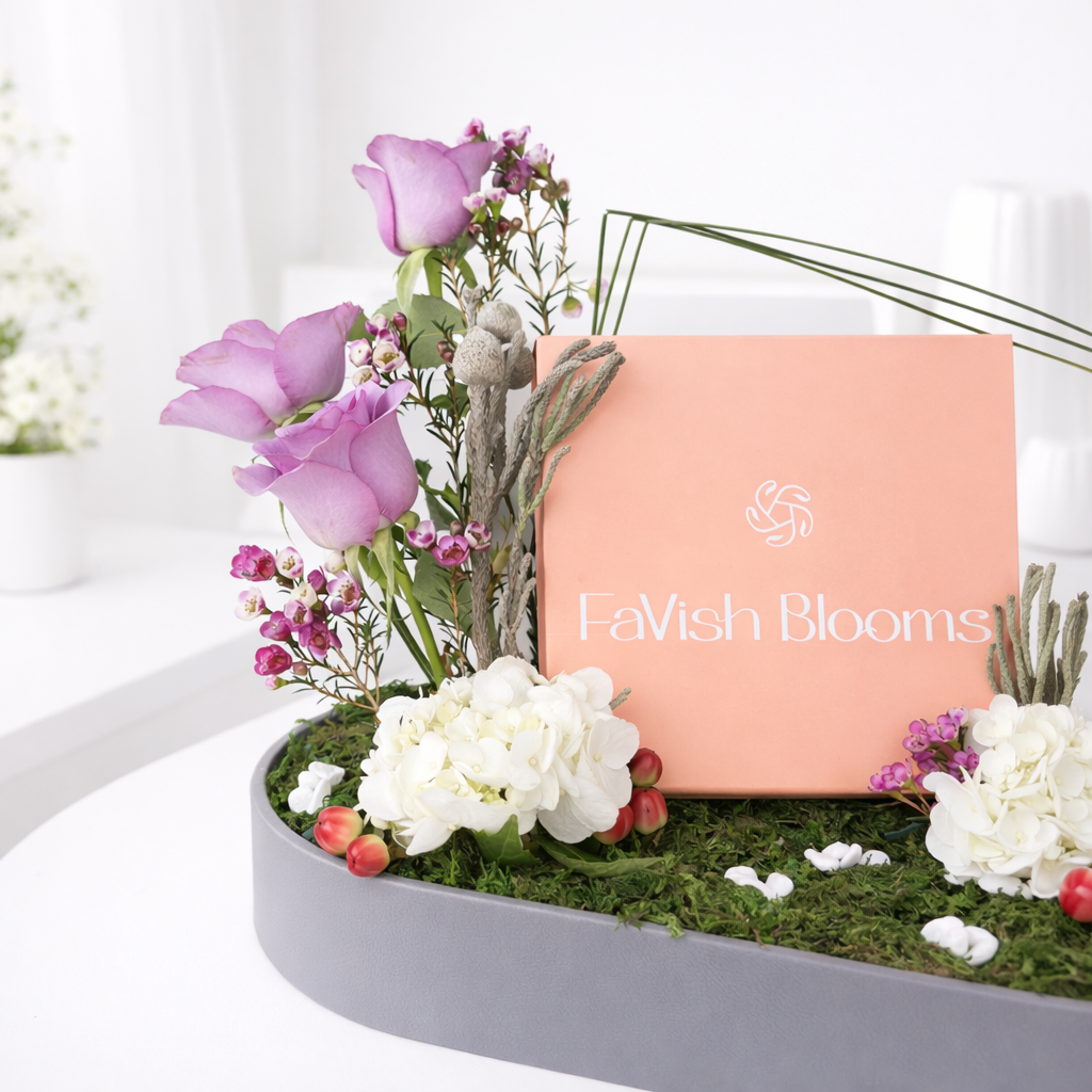 Close-up of a FaVish Blooms branded card nestled among purple roses, white hydrangeas, wax flowers, and dried botanicals on a moss-covered tray.