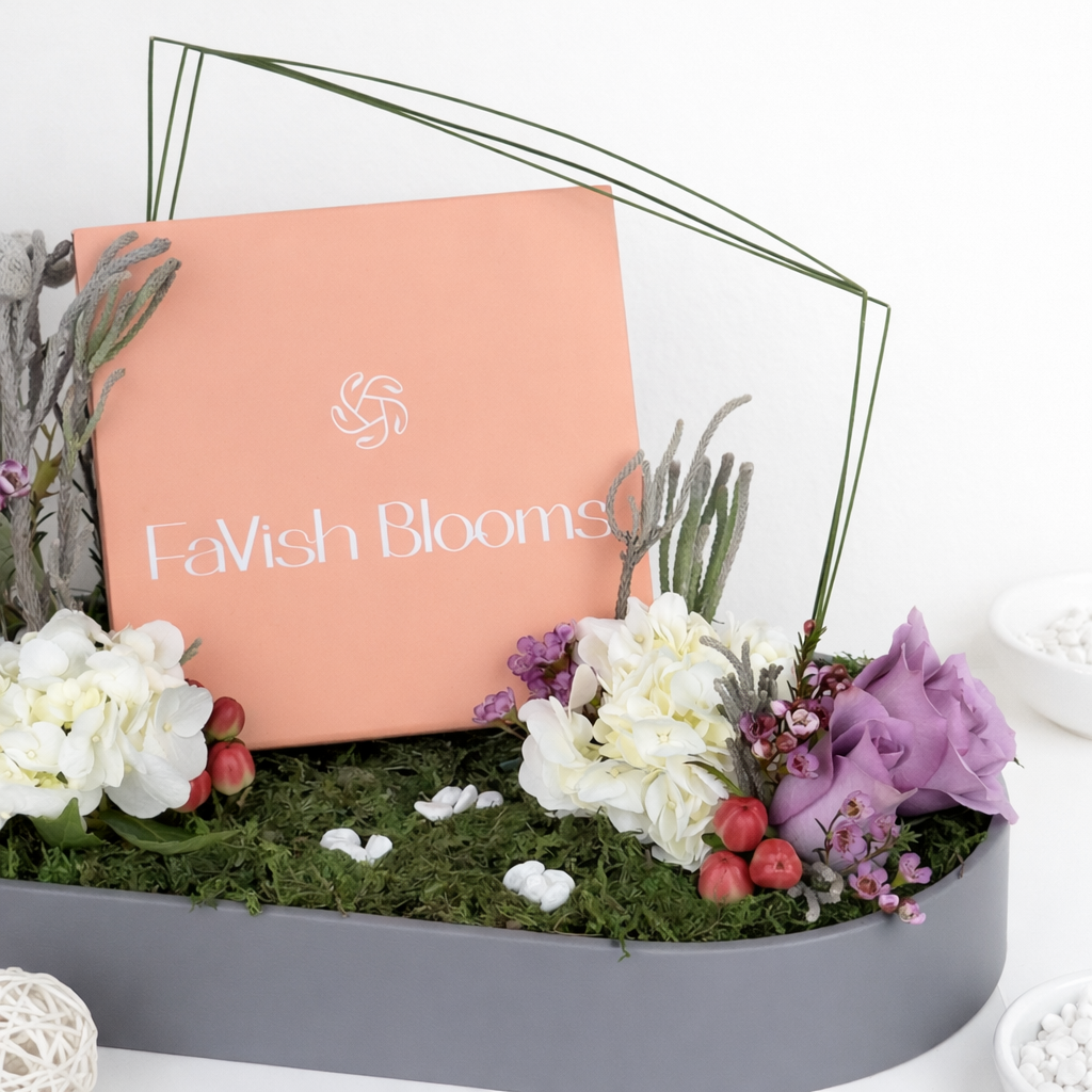 Close-up of a FaVish Blooms peach card with geometric wire accent surrounded by white hydrangeas, purple roses, and dried botanicals on a moss tray.