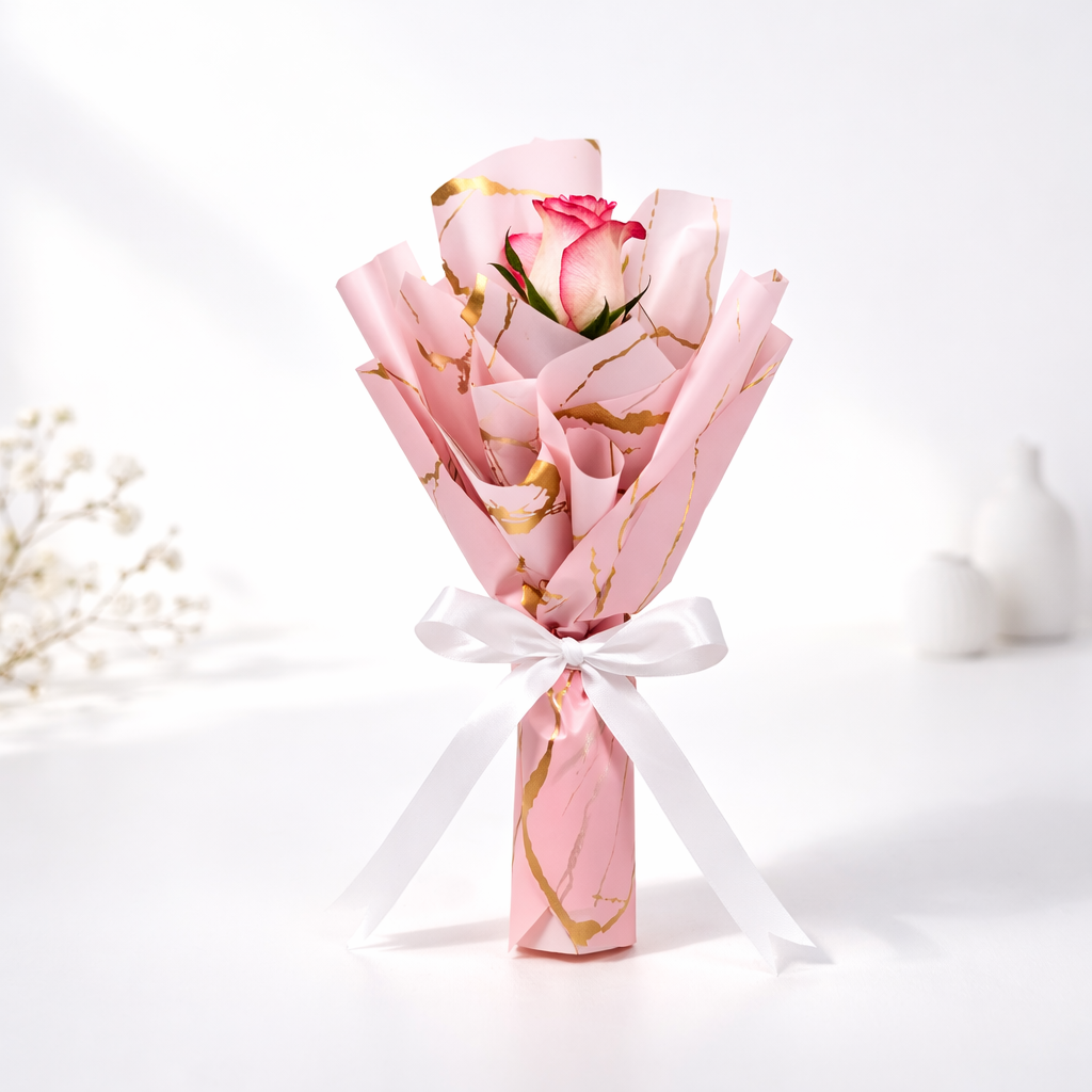 Single pink rose wrapped in pink and gold marble print paper tied with a white satin ribbon.