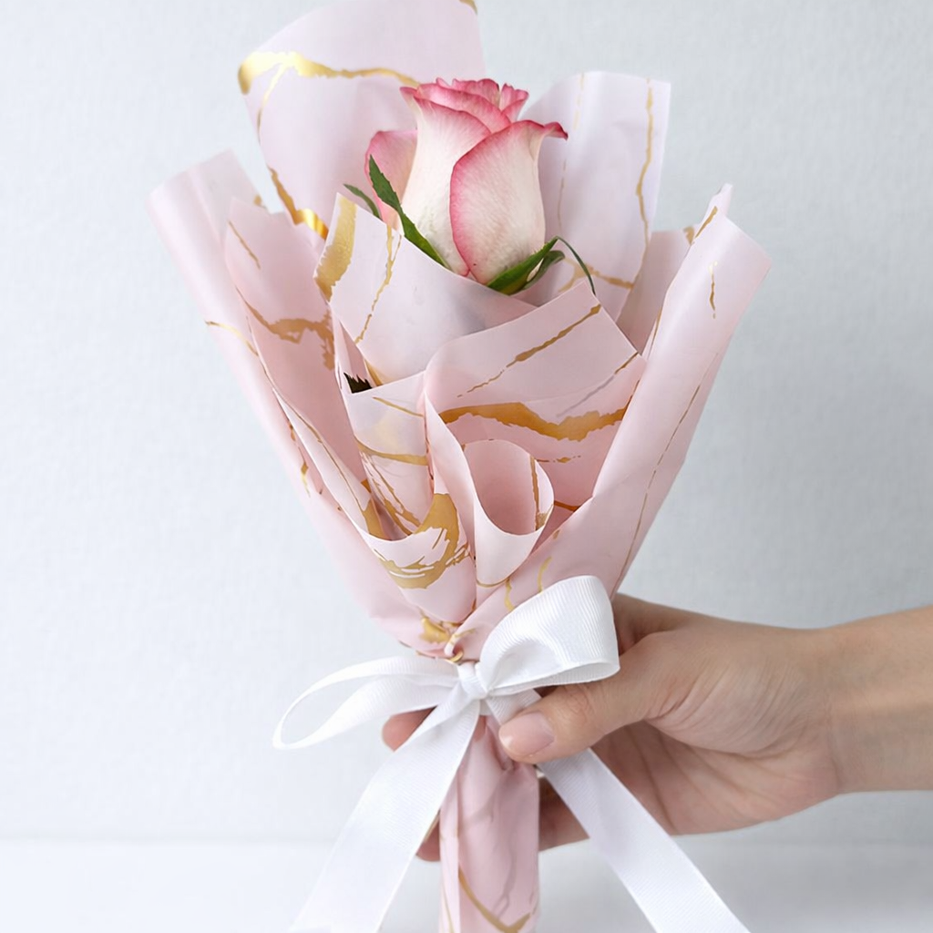Hand holding a single pink rose wrapped in pink and gold marble print paper with a white ribbon bow.