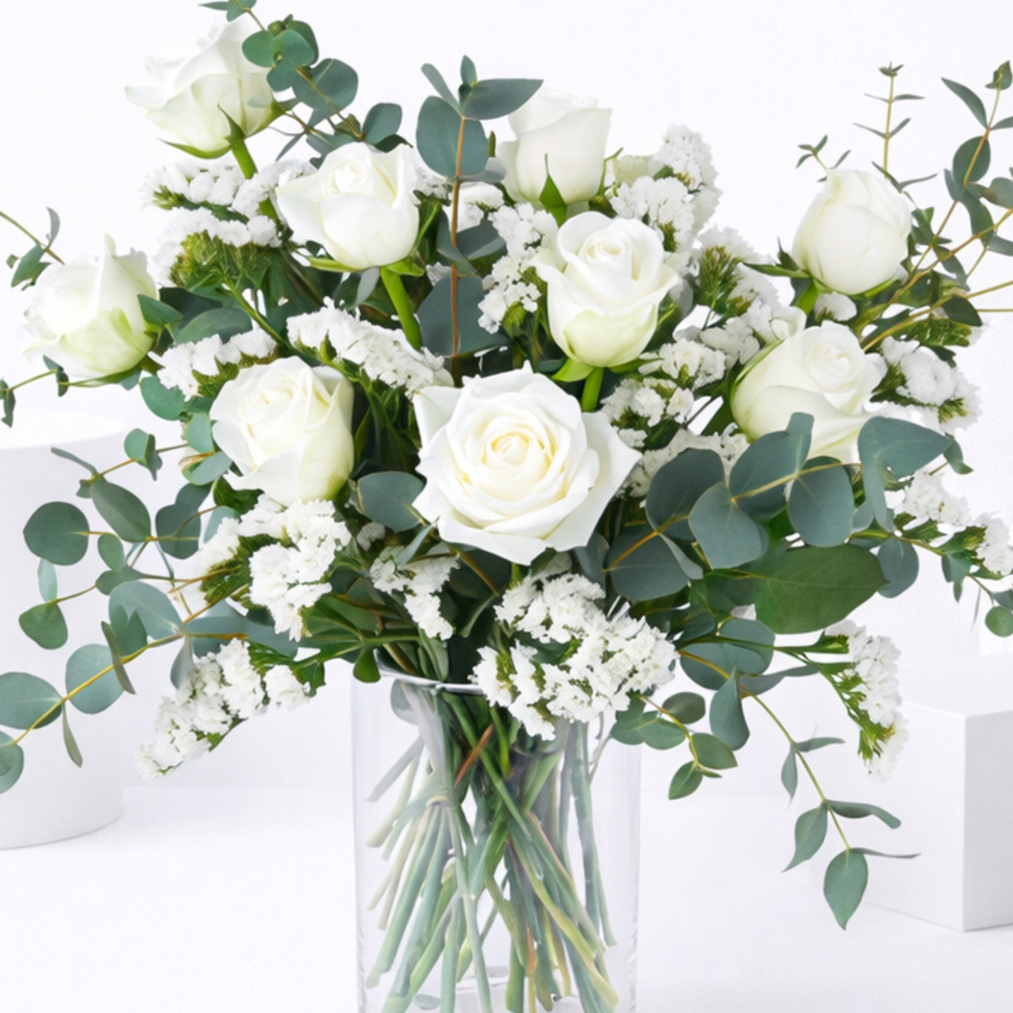 A glass vase of white roses with white statice and eucalyptus branches on a white surface.