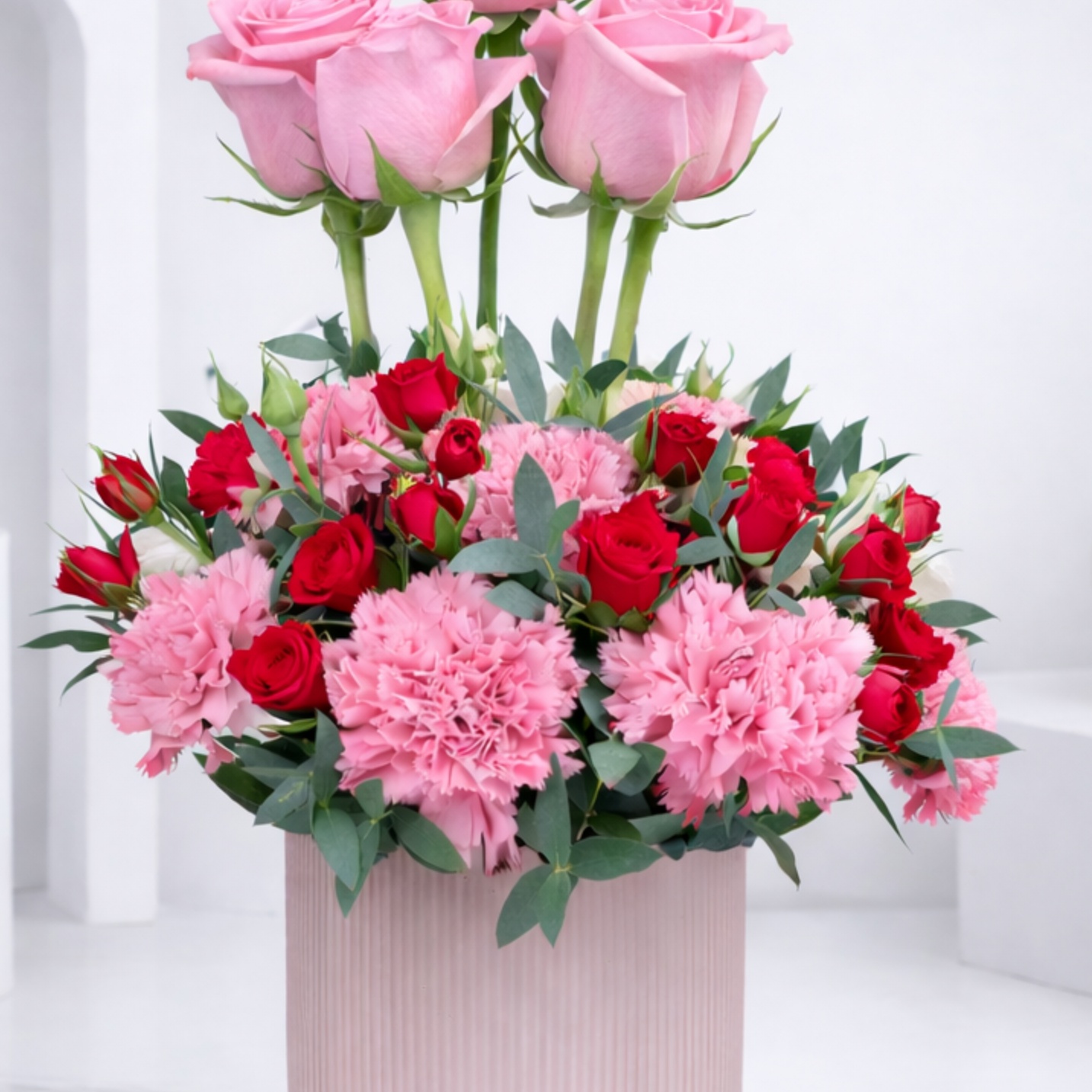 A pink gift box arrangement of tall pink roses, pink carnations, and red spray roses with green foliage.