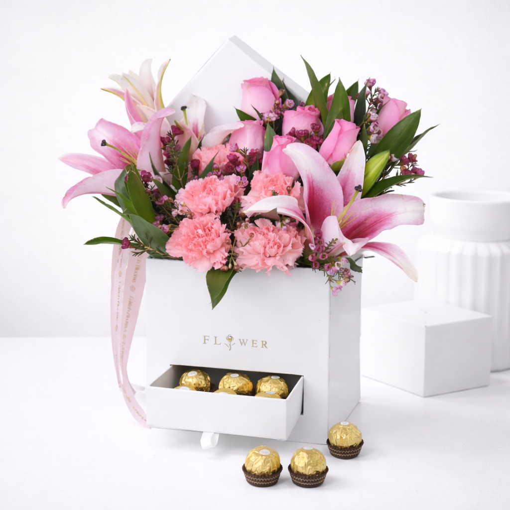 Pink lilies, roses, and carnations with wax flowers in a white square flower box with a drawer holding Ferrero Rocher chocolates.