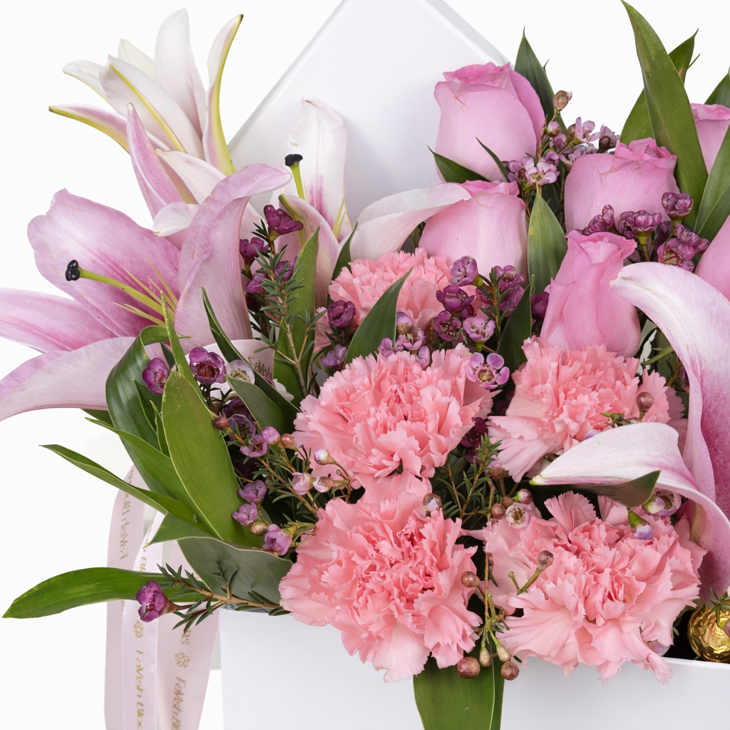 Macro shot of pink lilies, carnations, roses, and purple wax flowers in a white flower box with a Favish Blooms ribbon.