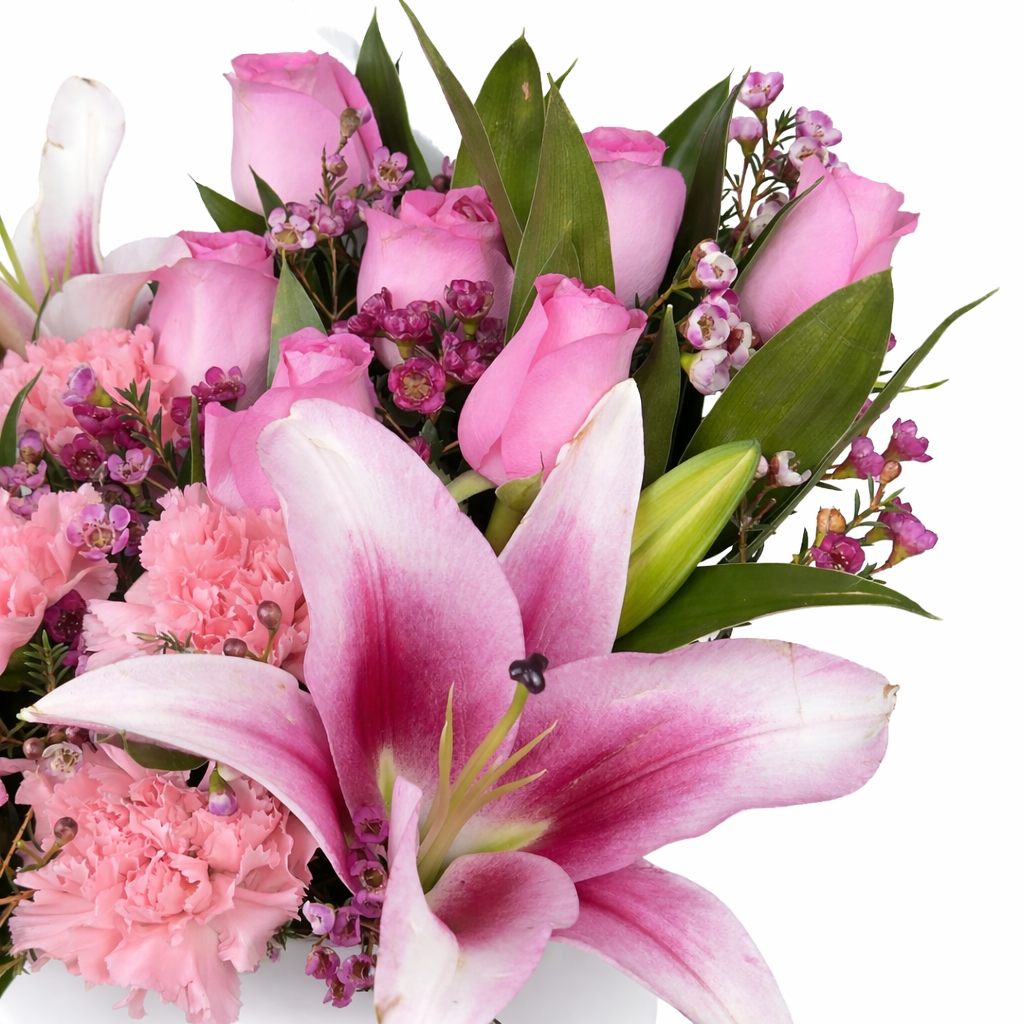 Close-up of a pink floral arrangement featuring pink roses, pink lilies, soft pink carnations, and green foliage on a white background.