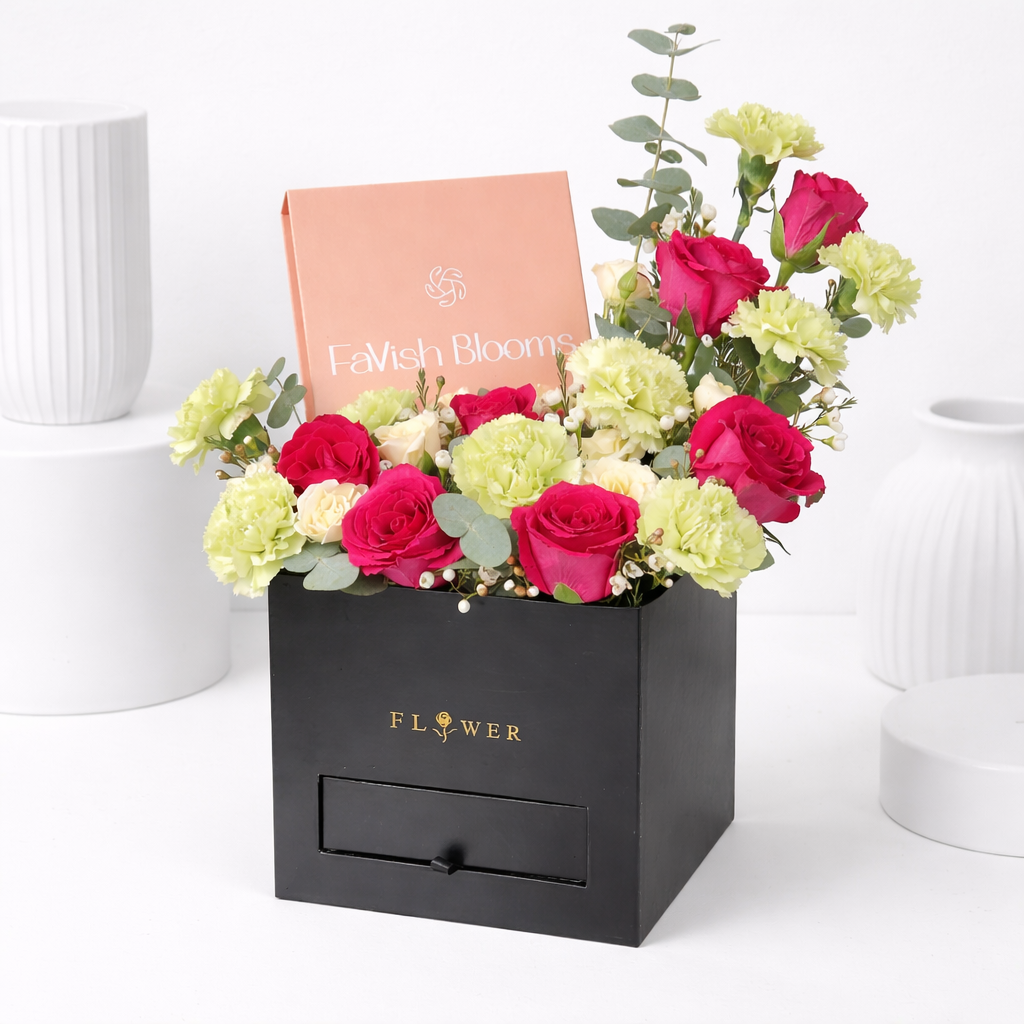 Hot pink roses, green carnations, and cream flowers with eucalyptus in a black square flower box with a drawer, featuring a Favish Blooms gift card.