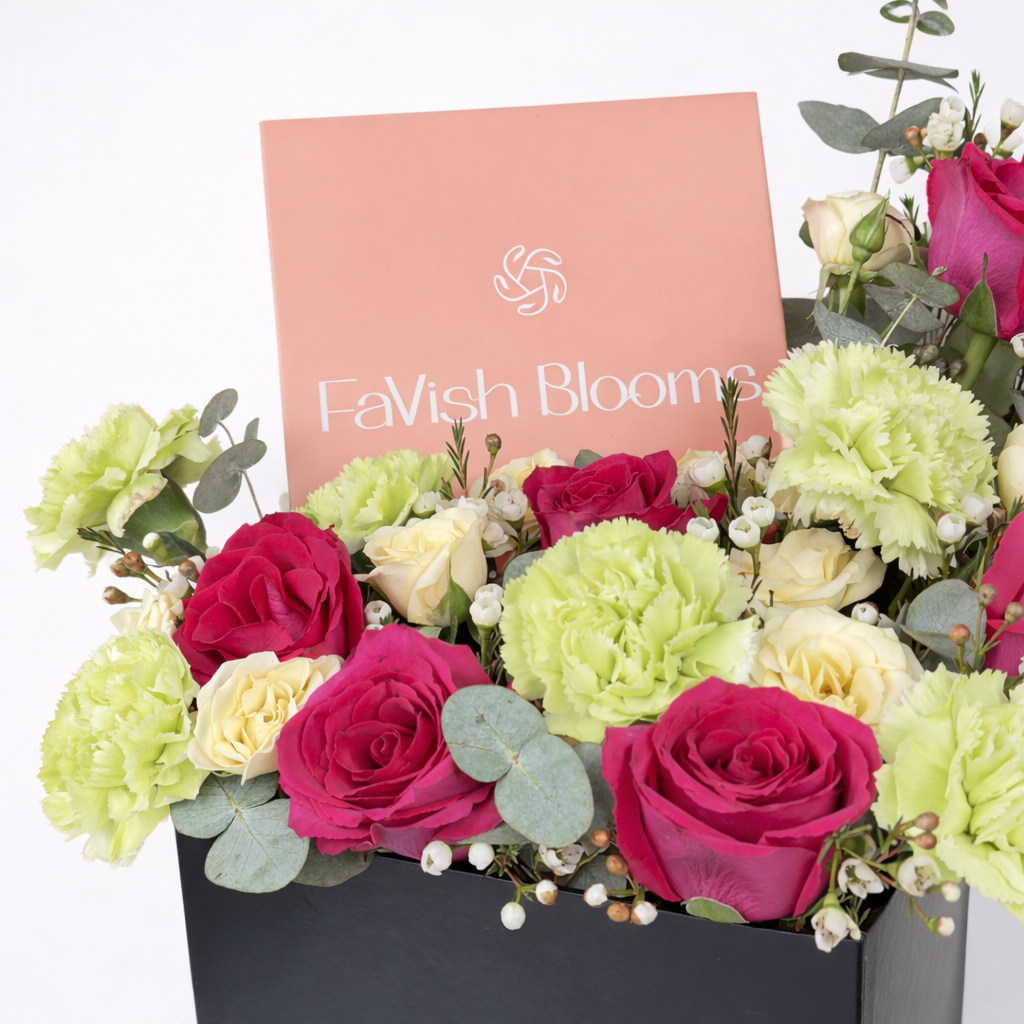 Close-up of hot pink roses, green carnations, cream spray roses, and white wax flowers with eucalyptus in a black box featuring a Favish Blooms gift card.