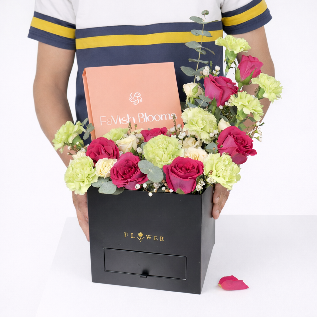 Person holding a black square flower box with a drawer filled with hot pink roses, green carnations, cream spray roses, and white wax flowers, featuring a Favish Blooms gift card.