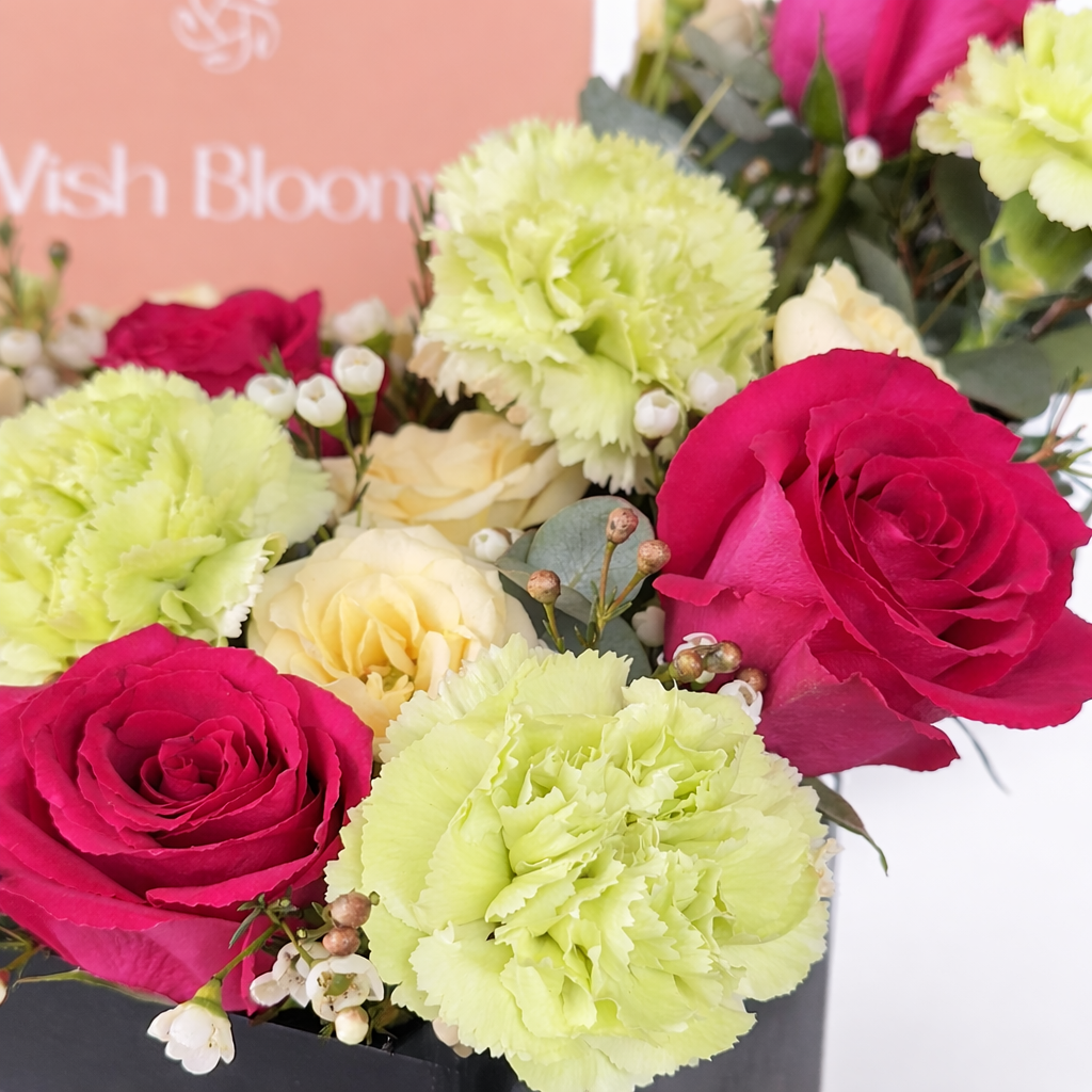 Macro shot of hot pink roses, green carnations, cream spray roses, and white wax flowers with eucalyptus in a black box with Favish Blooms branding in the background.