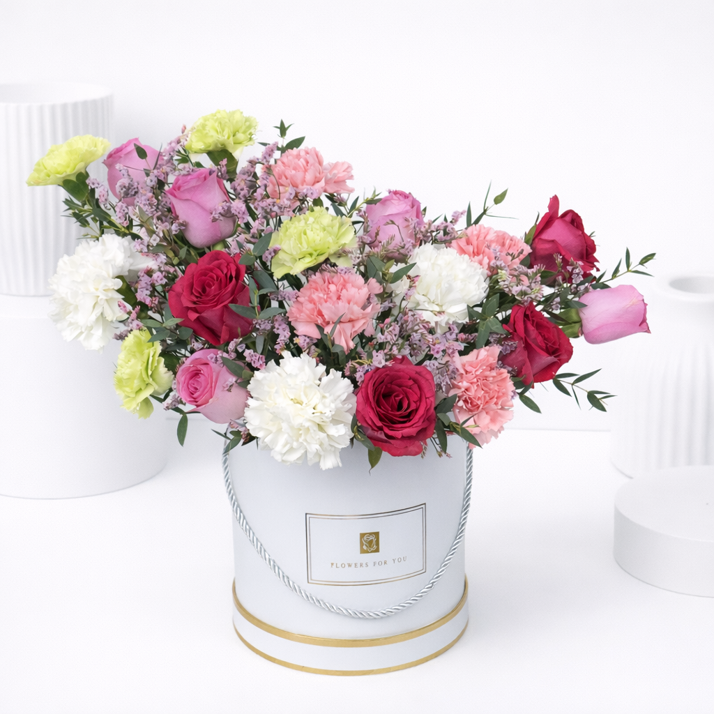 Red, pink, and lavender roses with white, pink, and green carnations and purple wax flowers in a white round gift box with gold trim.