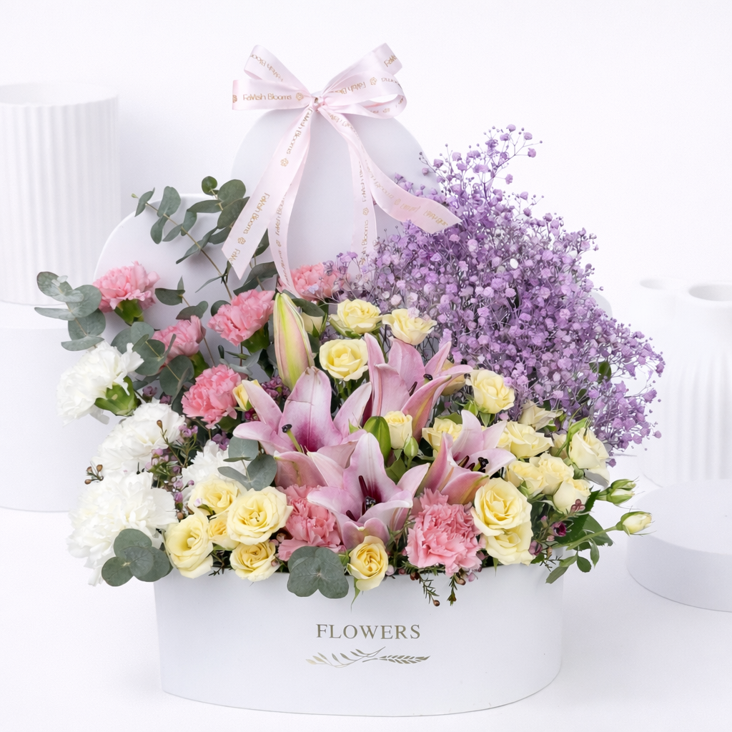 Pink lilies, cream roses, pink carnations, white flowers, and purple baby's breath arranged in a white oval flower box with a pink Favish Blooms ribbon bow.