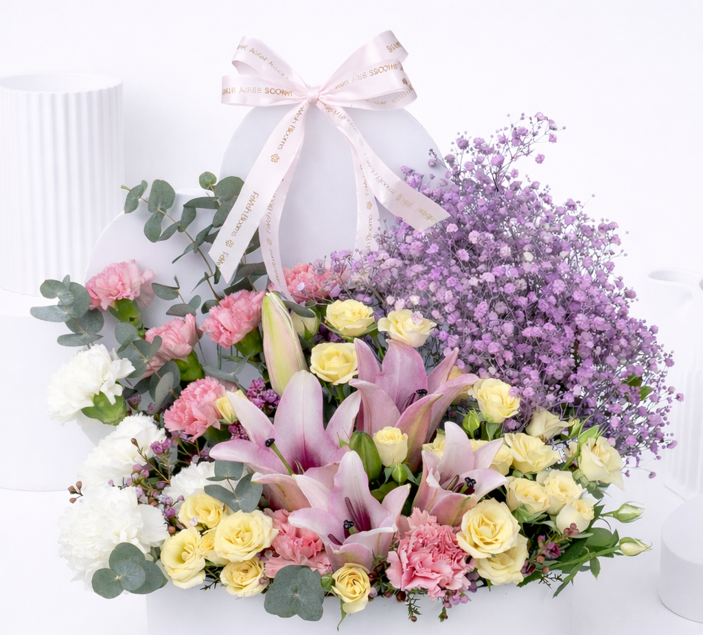 Close-up of pink lilies, cream roses, pink carnations, white flowers, and purple baby's breath in a white oval flower box with a Favish Blooms ribbon bow.