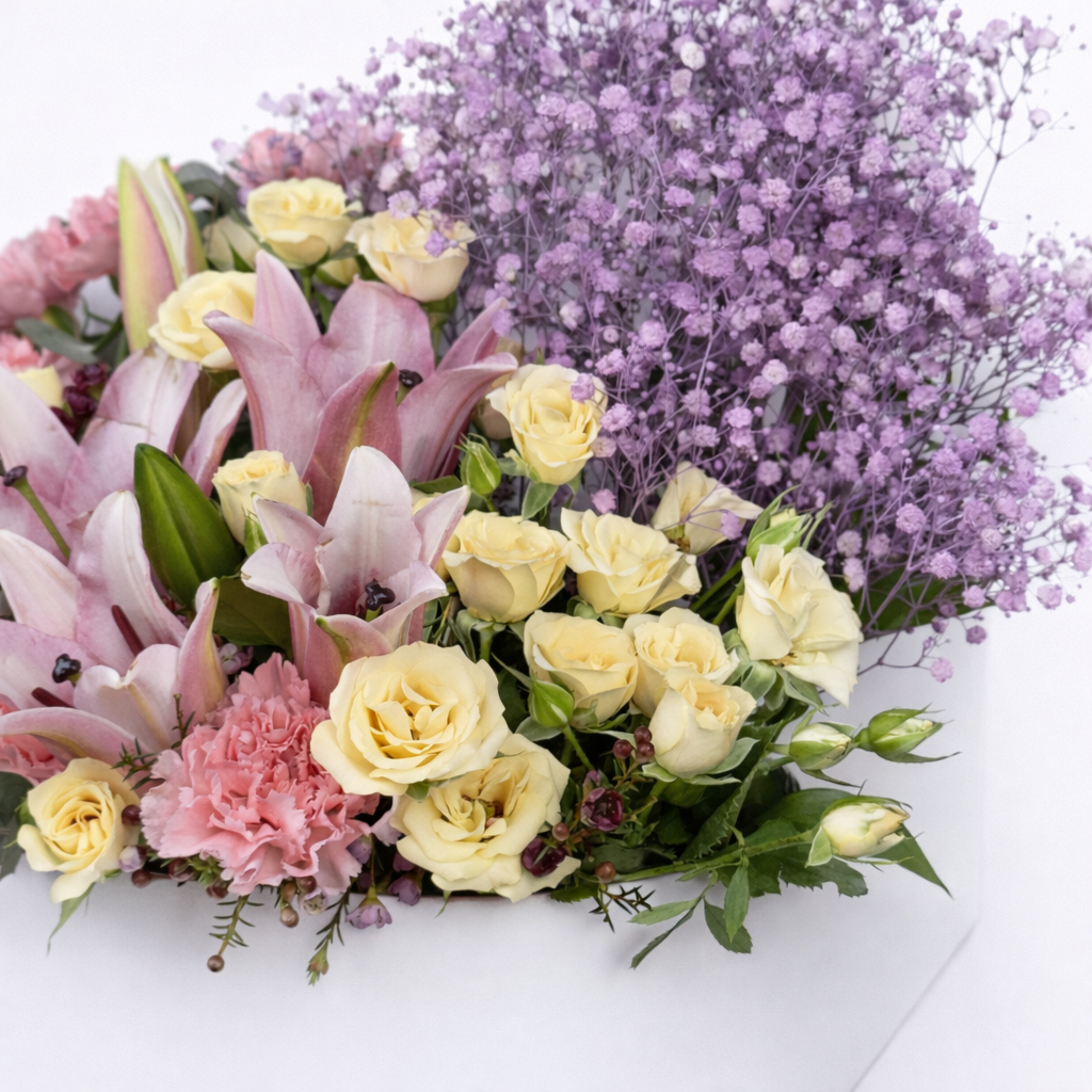 Detail shot of pink lilies, cream spray roses, pink carnations, and purple baby's breath in a white flower box.