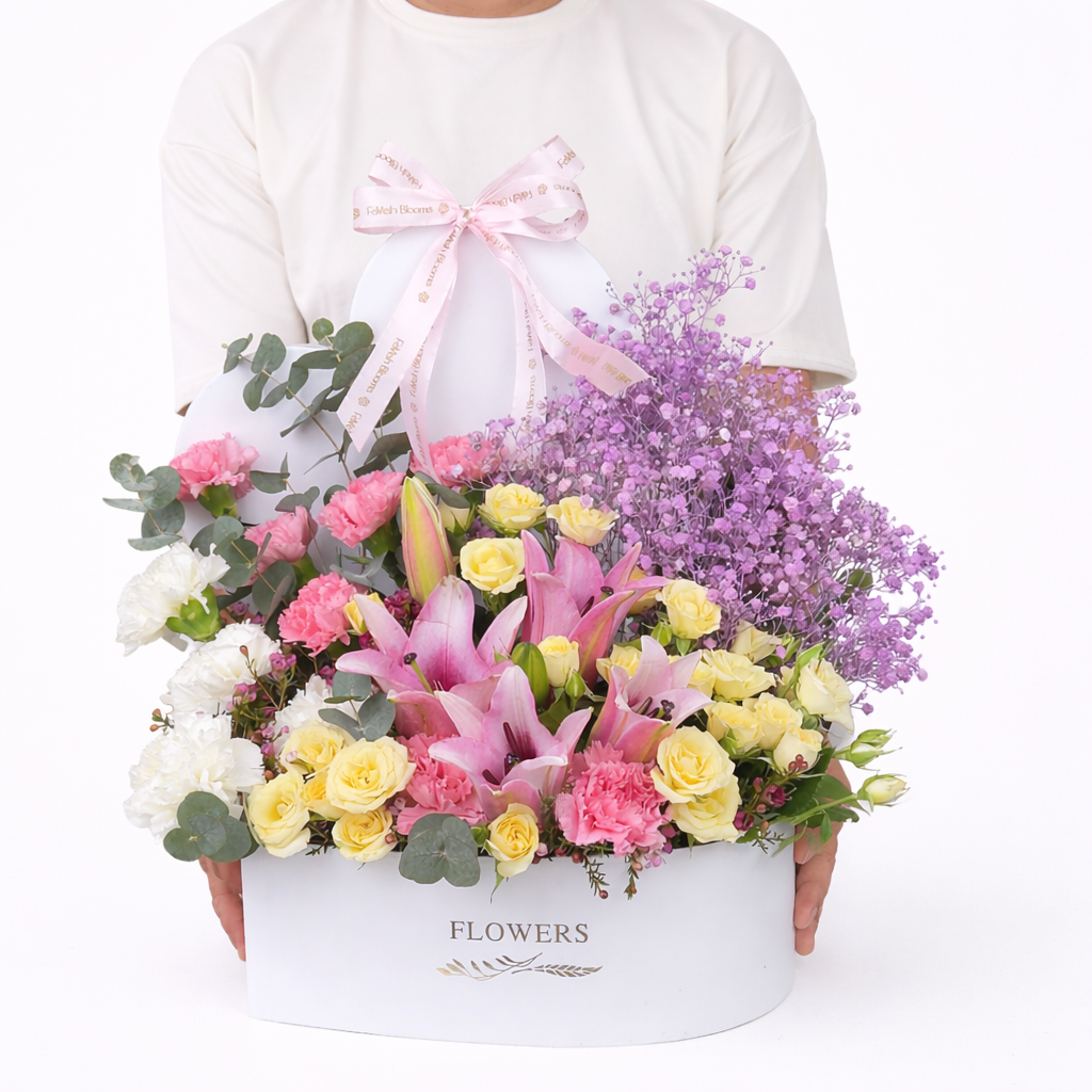 Person holding a large white oval flower box filled with pink lilies, cream roses, pink carnations, white flowers, and purple baby's breath with a Favish Blooms ribbon bow.