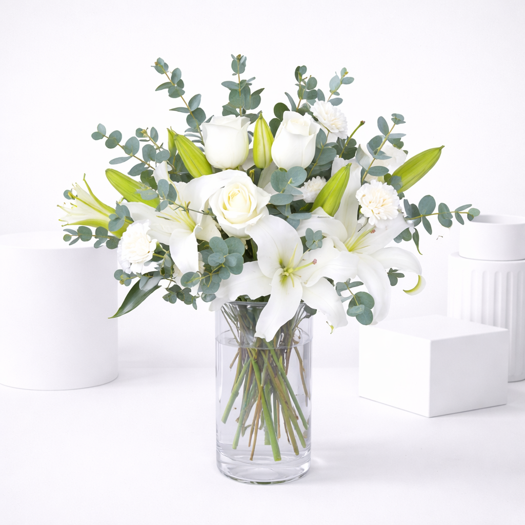 White lilies, roses, and carnations with eucalyptus arranged in a clear glass vase.