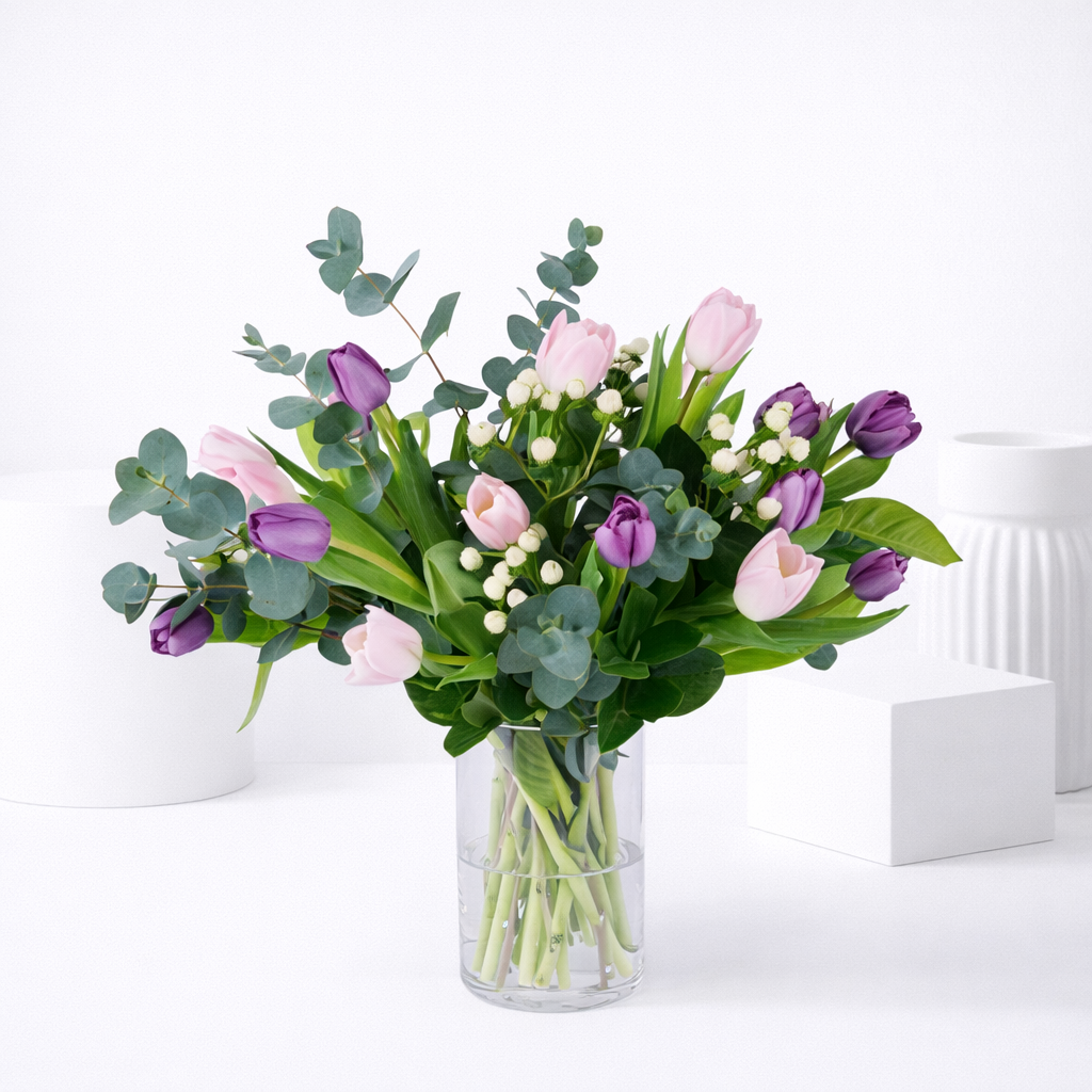 Pink and purple tulips with white berry stems and eucalyptus arranged in a clear glass vase.