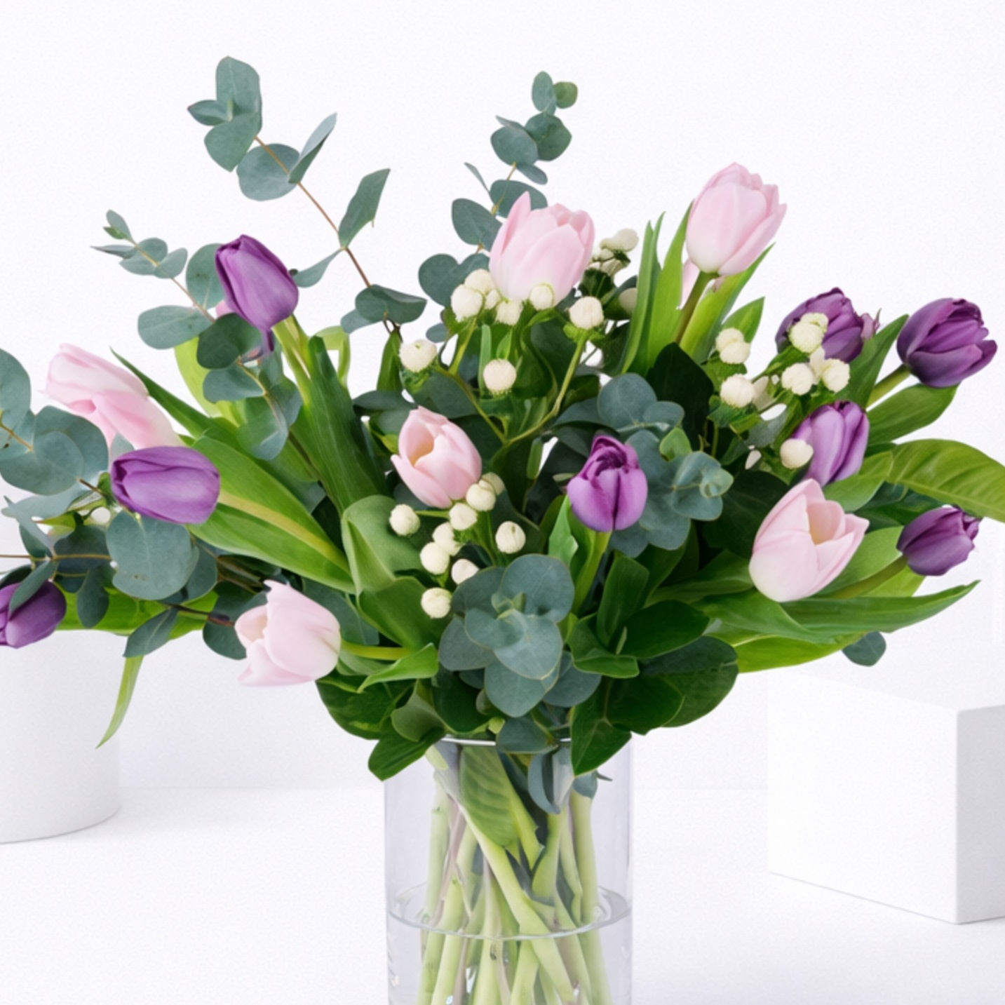 A glass vase of pink and purple tulips with white berry stems and eucalyptus foliage on a white surface.