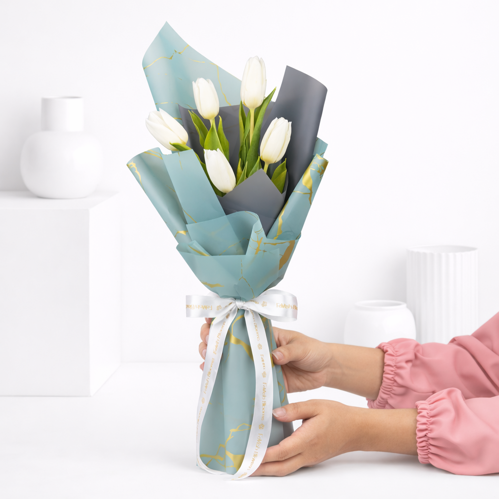 Person in a pink top holding a white tulip bouquet wrapped in blue-grey and gold-print paper with a white Favish Blooms ribbon.
