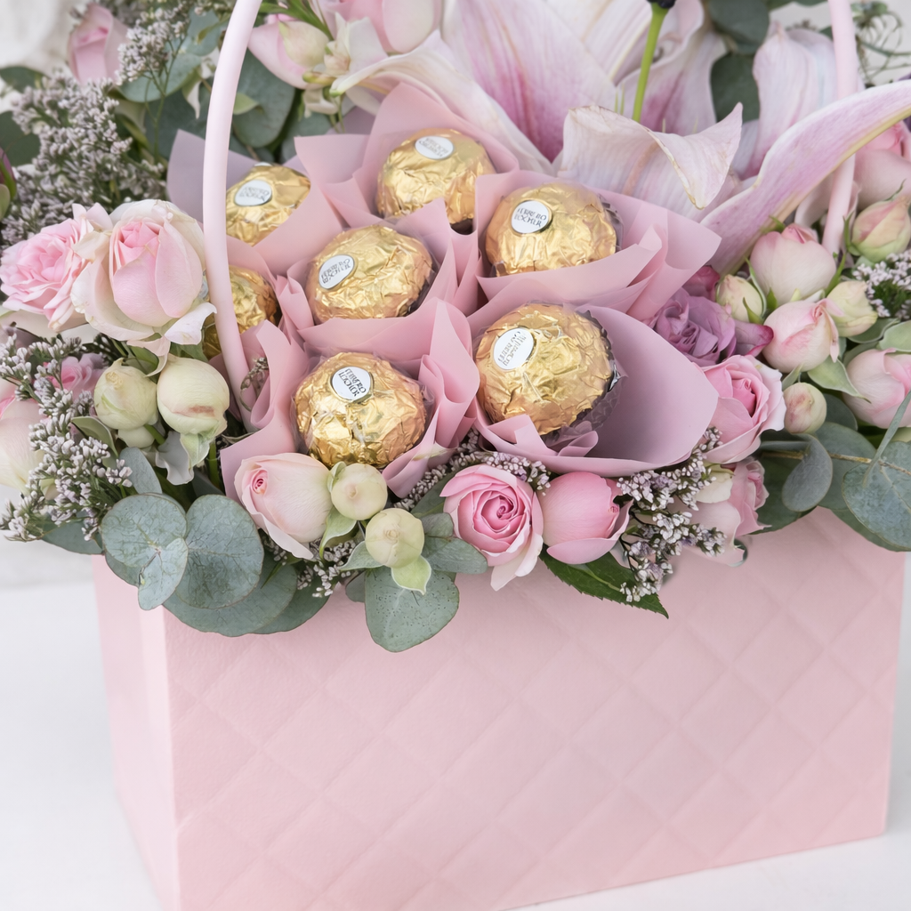 Close-up of Ferrero Rocher chocolates nestled among pink spray roses, lilies, and eucalyptus in a pink quilted handbag-style flower box.