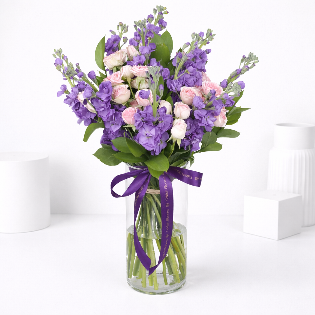 Purple stock flowers and pink spray roses in a glass vase tied with a purple ribbon.