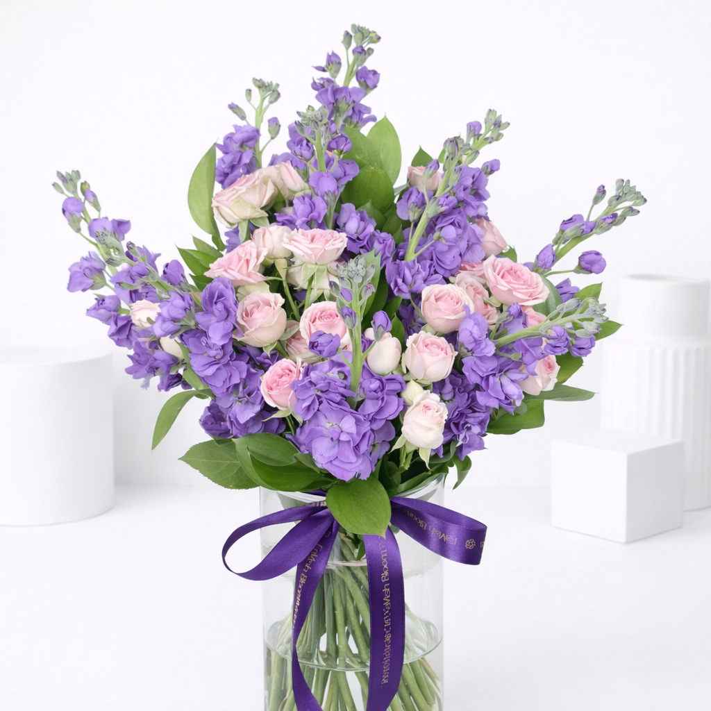 Purple stock flowers and pink spray roses in a glass vase with purple branded ribbon.