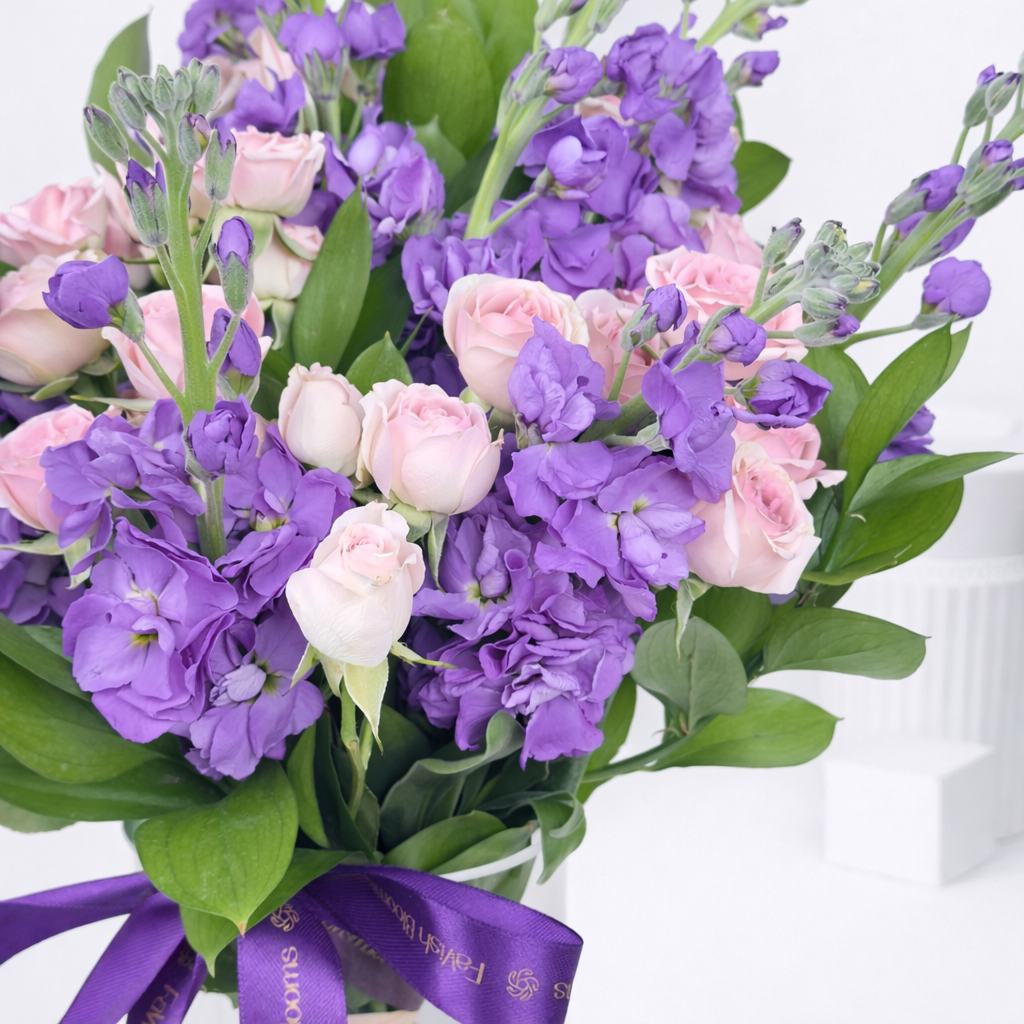 Close-up of purple stock flowers and pink spray rose buds with purple branded ribbon.