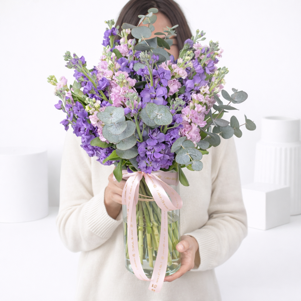 "Woman in white sweater holding glass vase of purple and pink stock flowers with eucalyptus"
