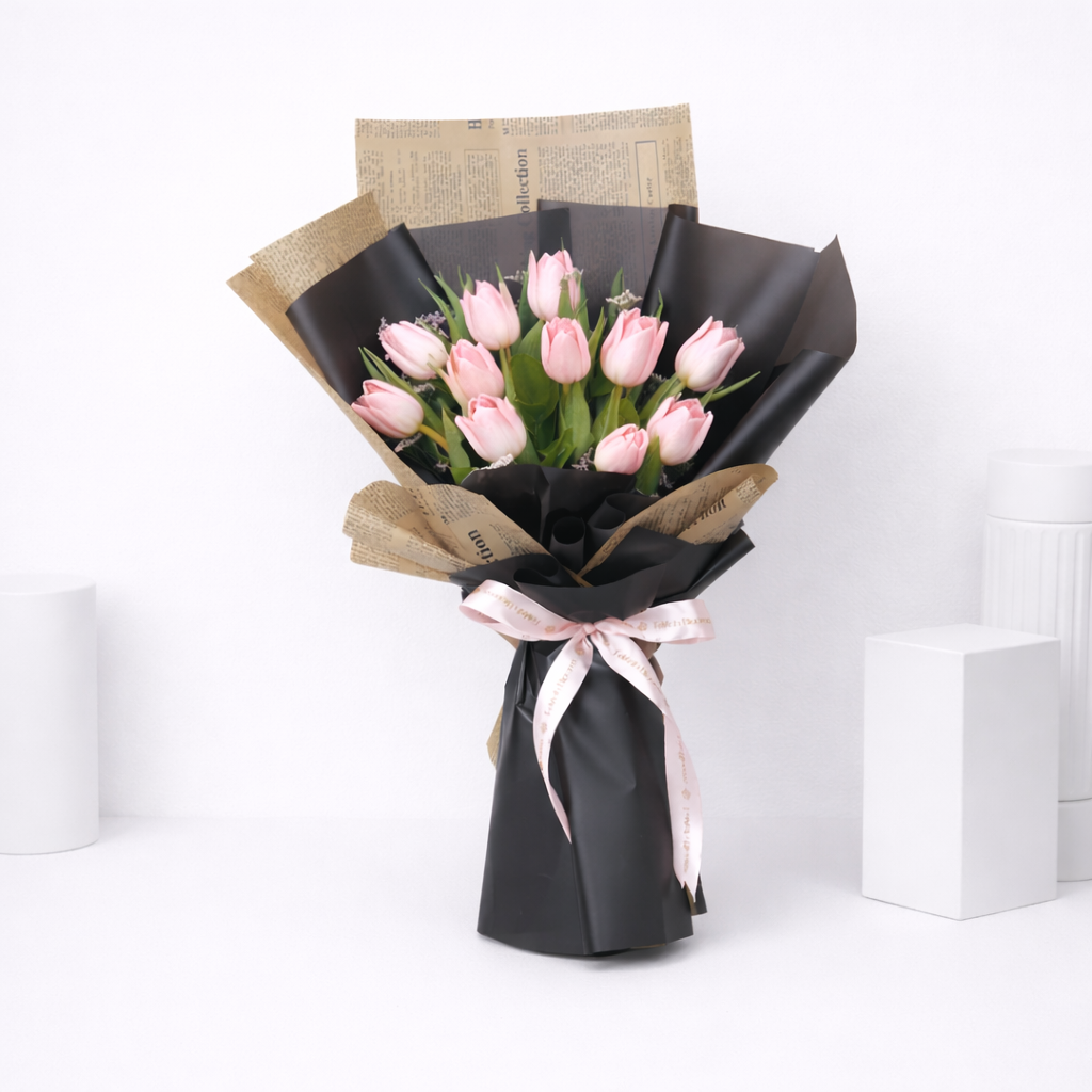 Pink tulip bouquet wrapped in black matte and newspaper kraft paper, tied with a pink satin ribbon, on a white background.