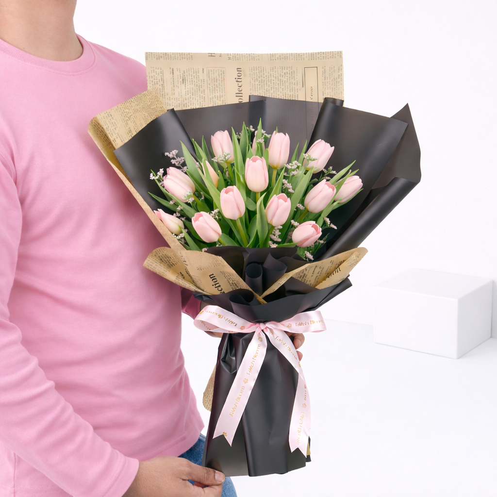A person holding a bouquet of pink tulips wrapped in black and newspaper-print paper, tied with a pink satin ribbon.