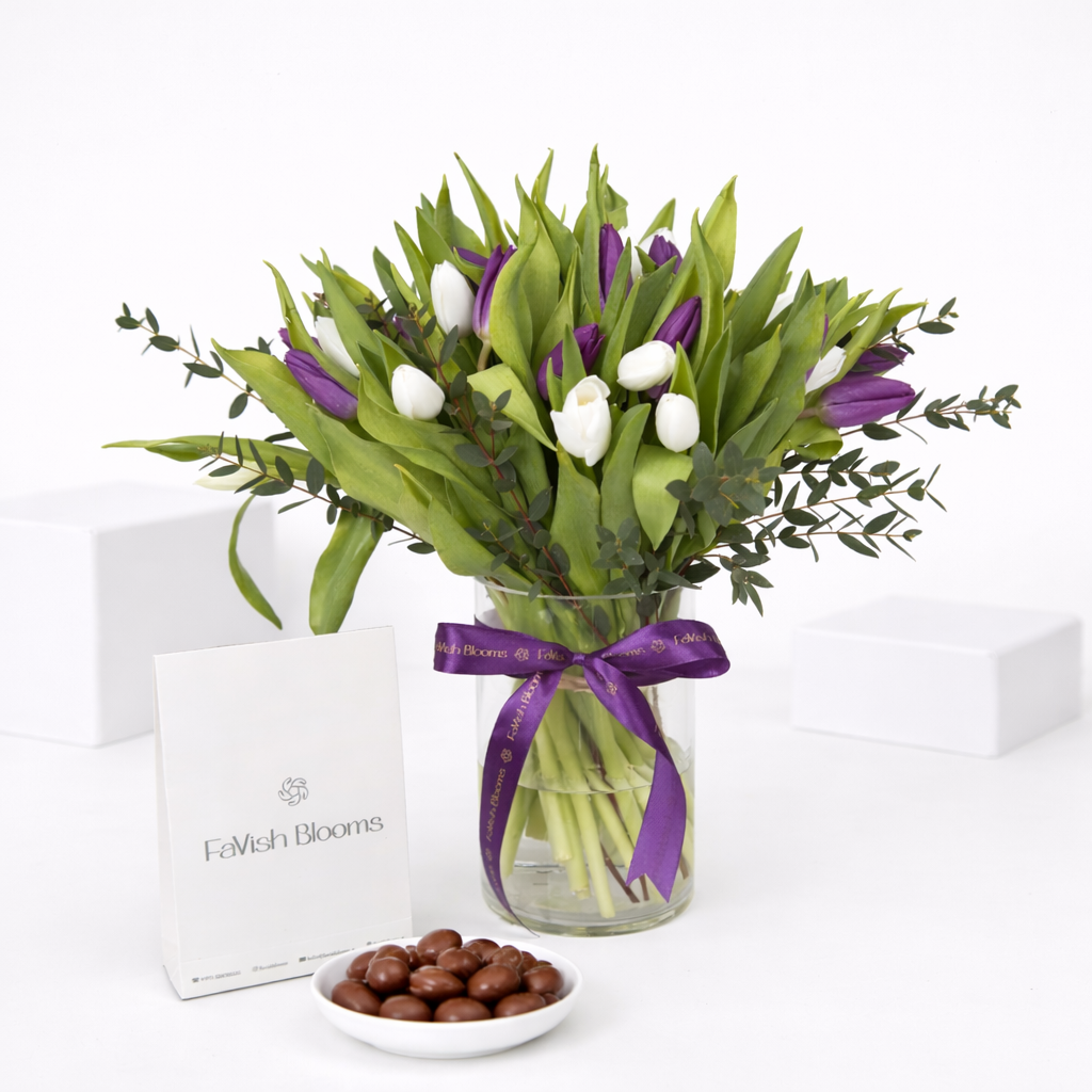 Purple and white tulips in a glass vase with a purple ribbon, beside chocolate-covered nuts and a FaVish Blooms card.