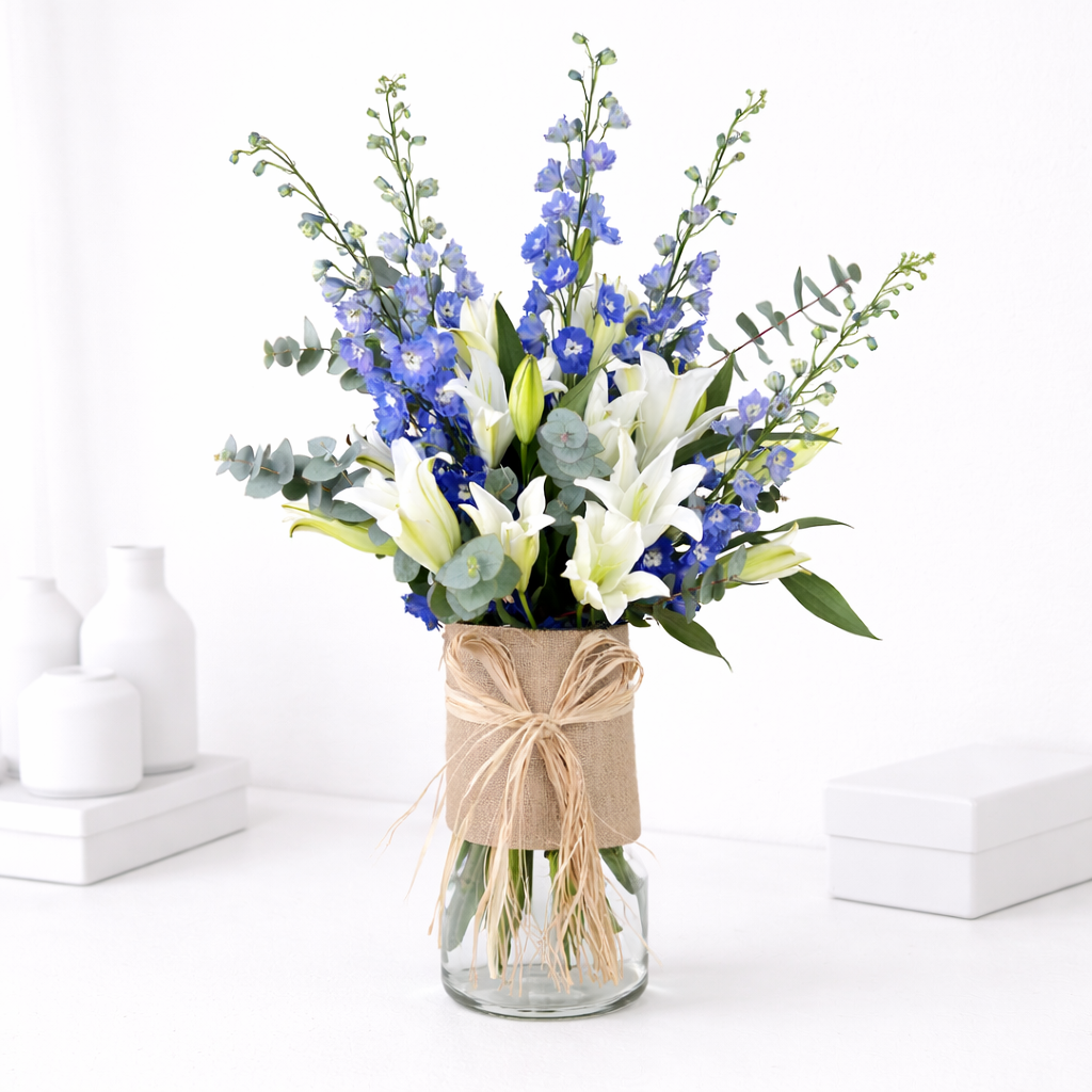 White lilies and blue delphinium with eucalyptus in a glass vase wrapped with burlap and raffia bow on a white surface.