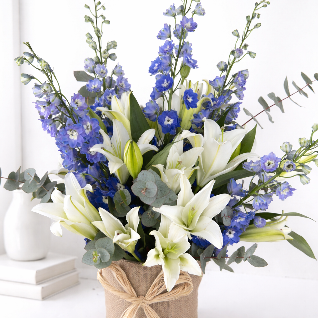 Close-up of white lilies and blue delphinium with eucalyptus in a burlap-wrapped vase with raffia bow.