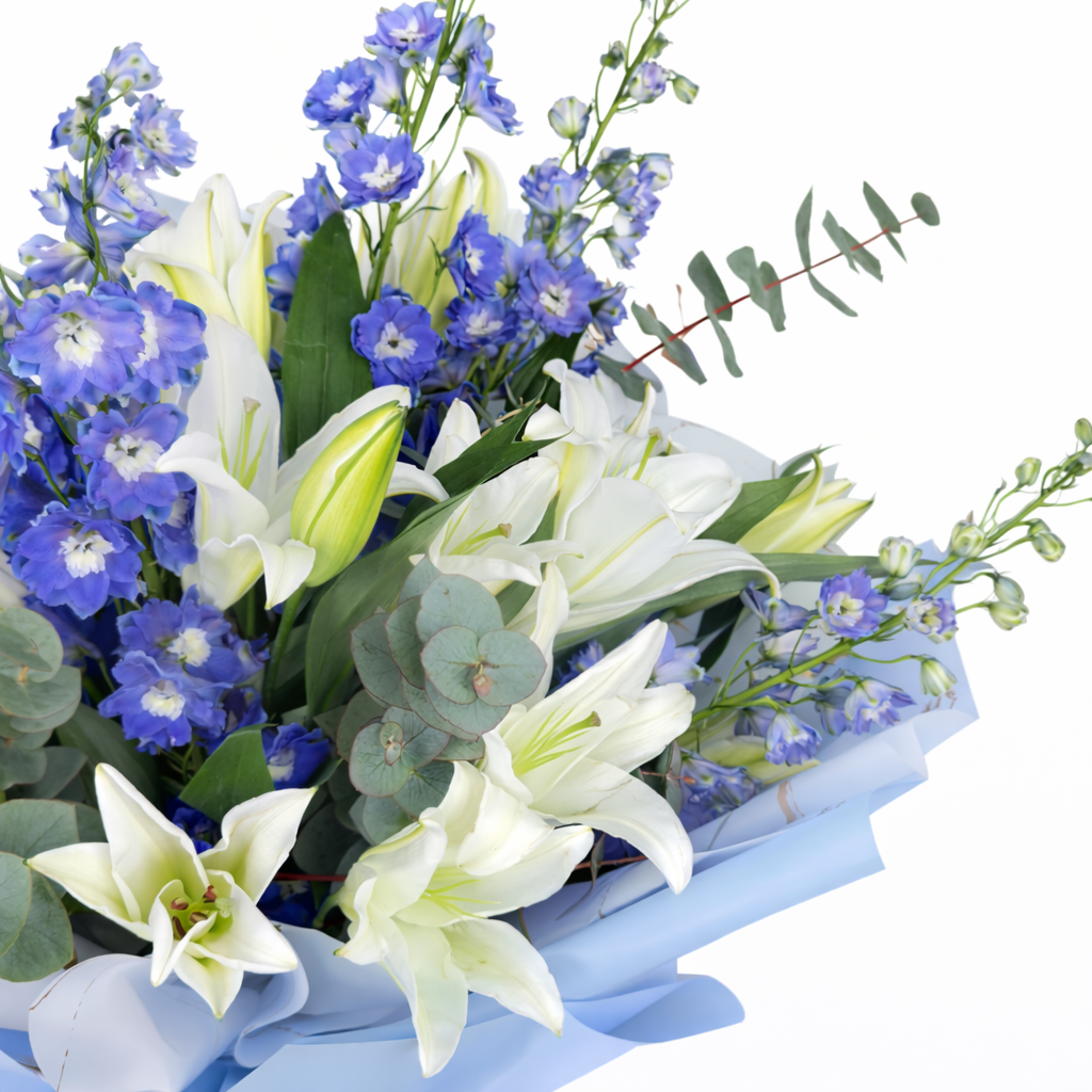 Close-up of open white lilies, blue delphinium buds, and eucalyptus in light blue wrapping.