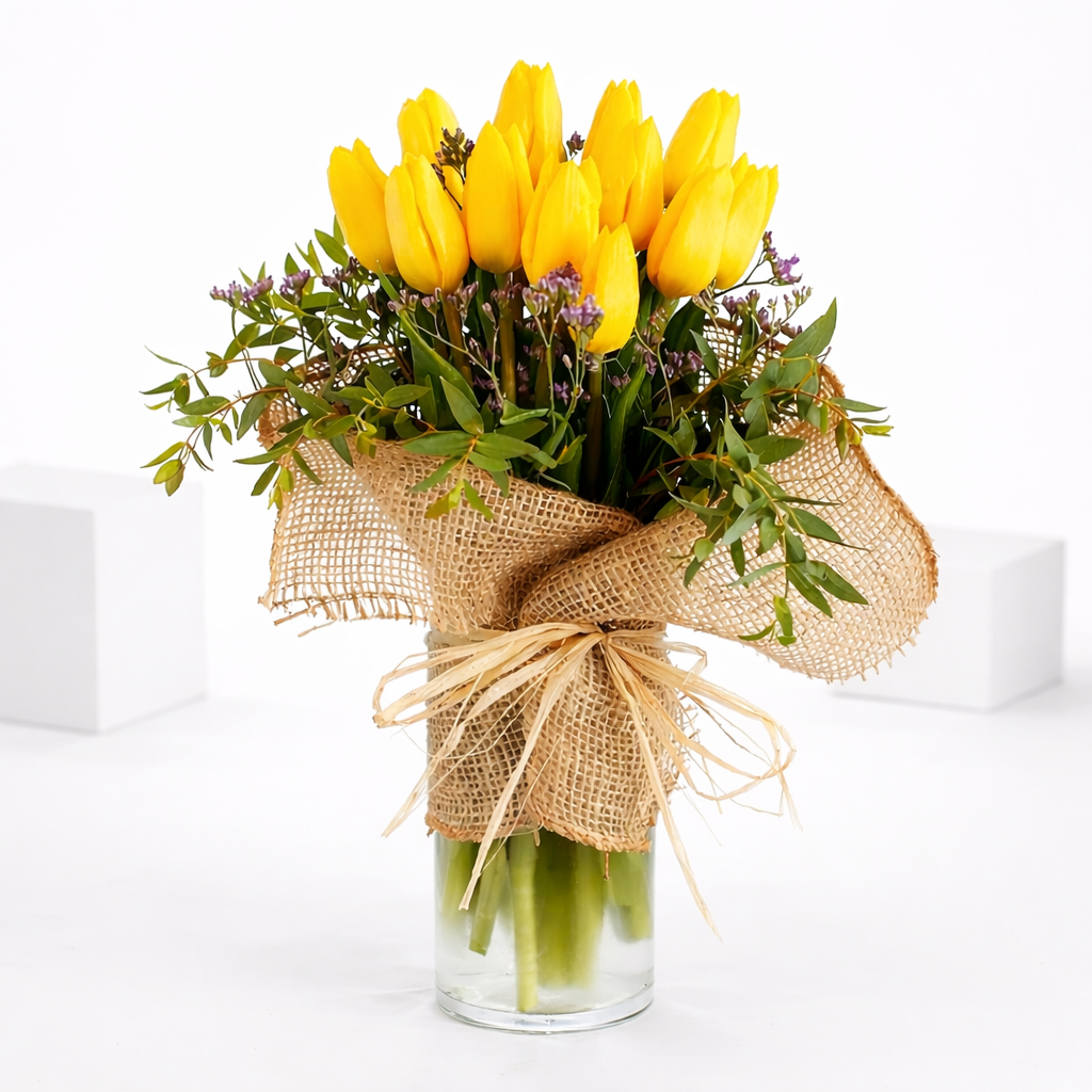 Yellow tulip bouquet with greenery in a glass vase wrapped in burlap and raffia bow.